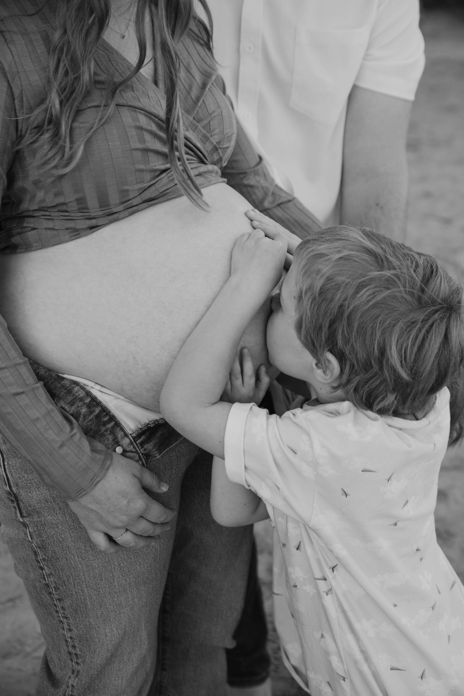 Black and white photo of young child kissing mothers pregnant belly outdoors.