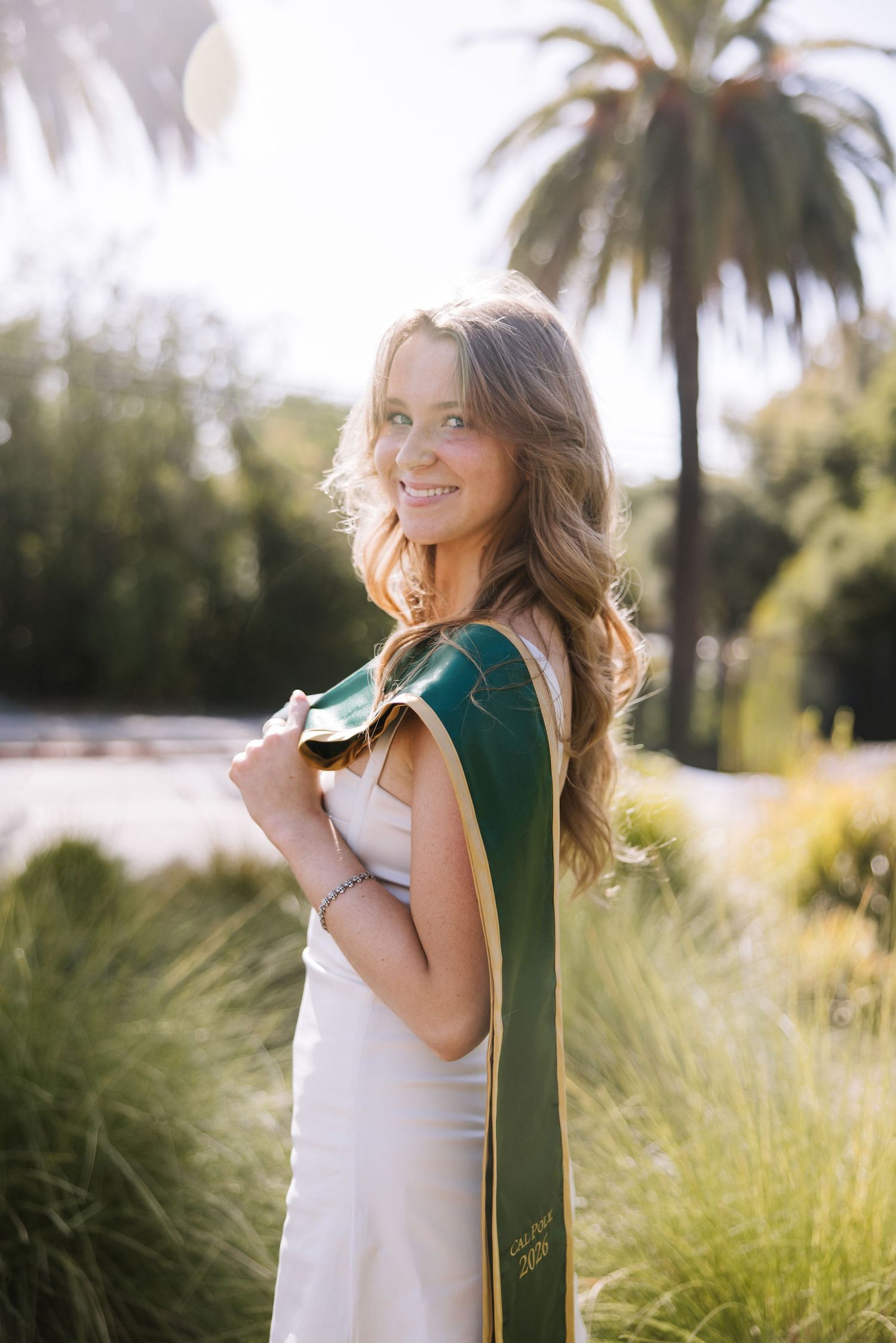 Smiling graduate in white dress poses with green Cal Poly 2024 sash near palm trees.