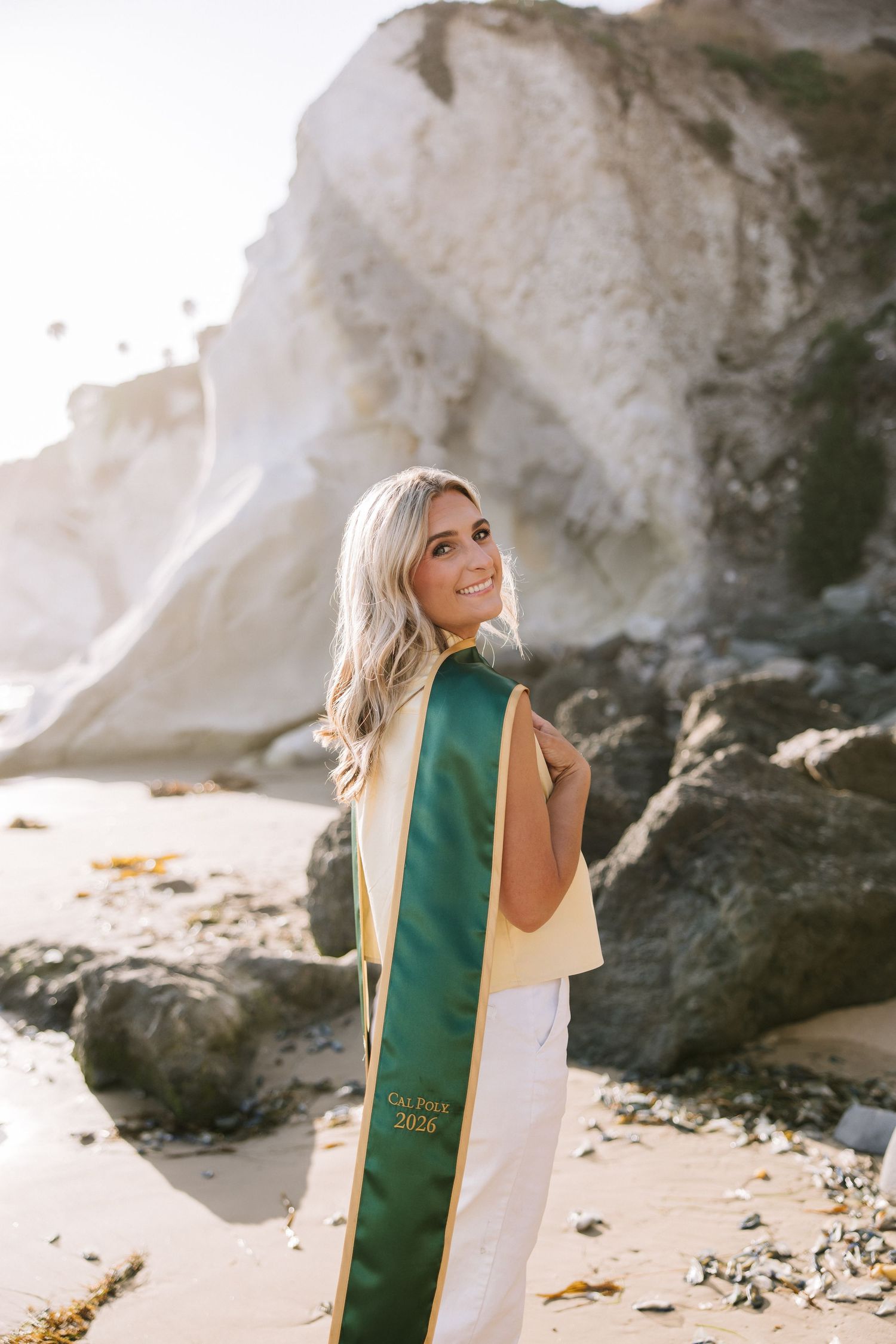 Blonde woman wearing Cal Poly 2026 green graduation stole smiling on rocky beach at golden hour.