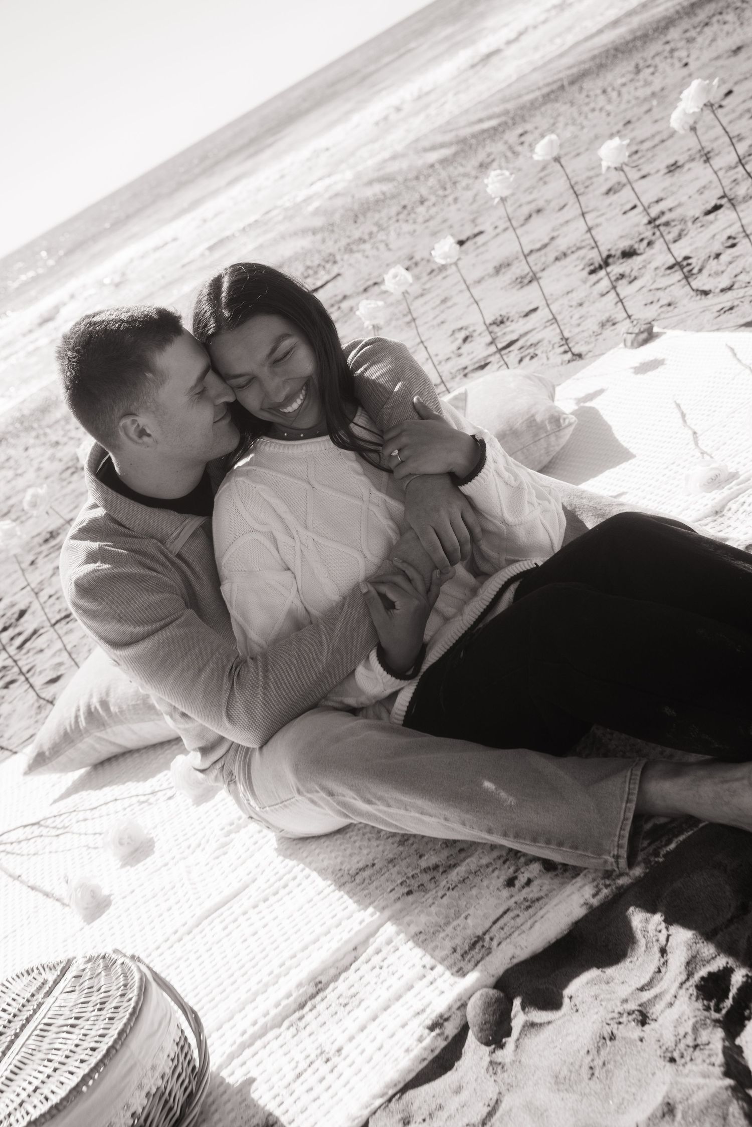 Black and white photo of a happy couple cuddling on a beach blanket after their engagement.