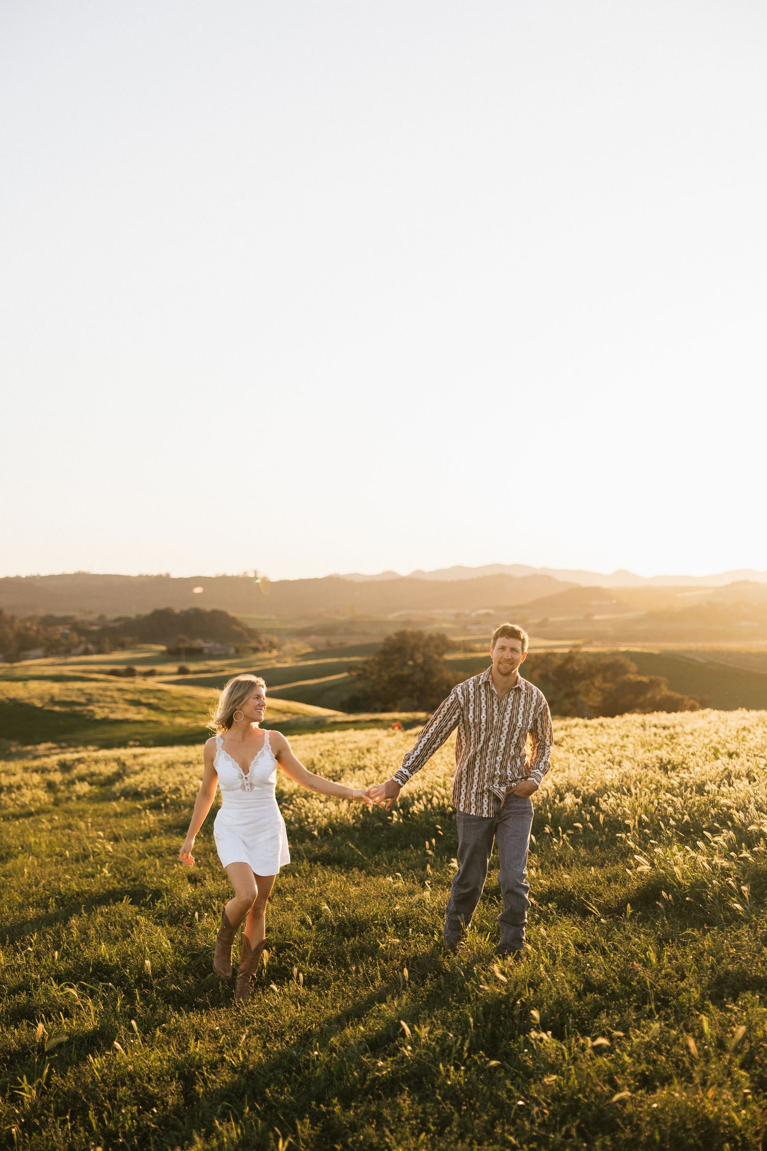 Couple holding hands walking through a green meadow with rolling hills at sunset.