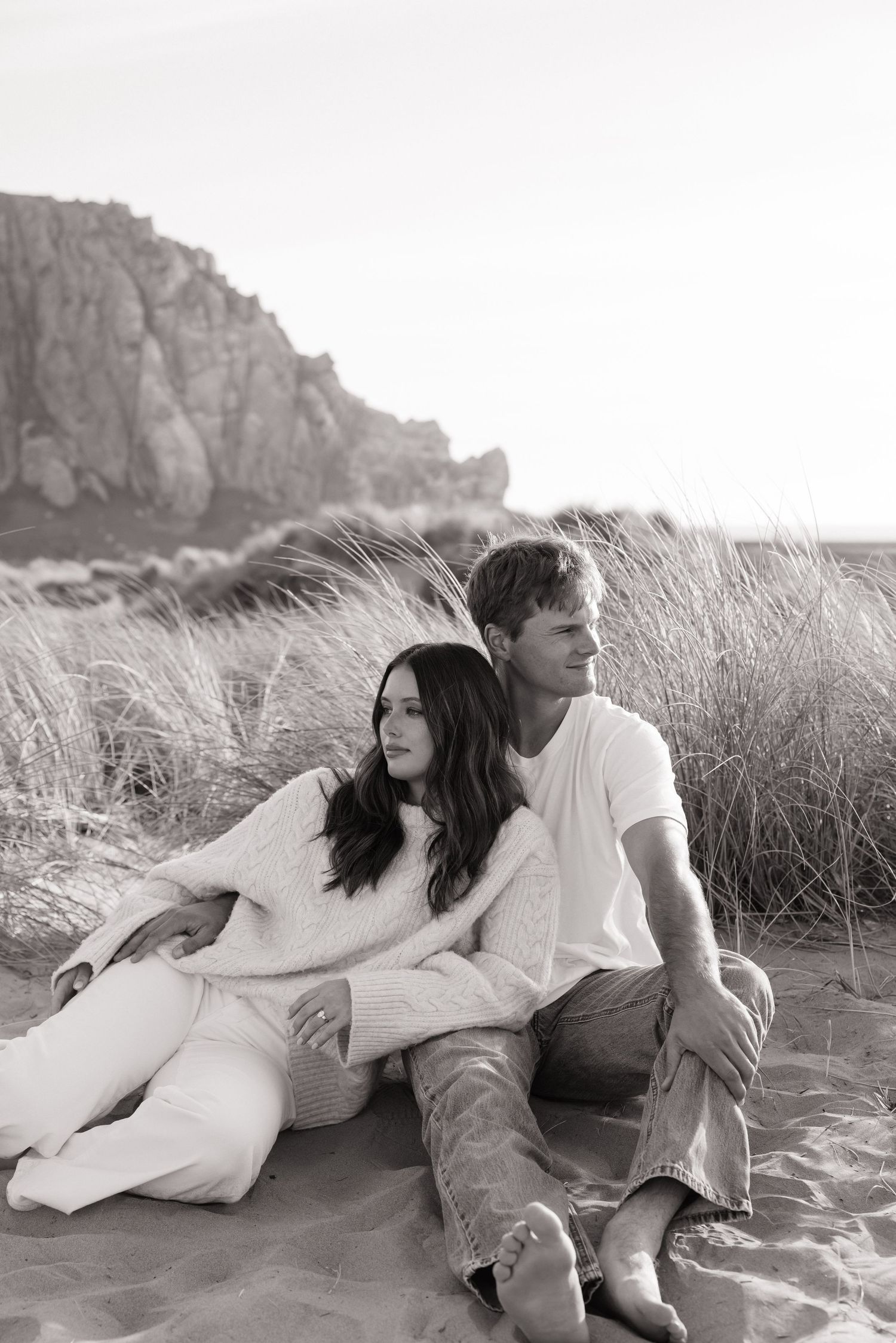 Couple sitting together on beach sand dunes with rocky cliffs in black and white photography.