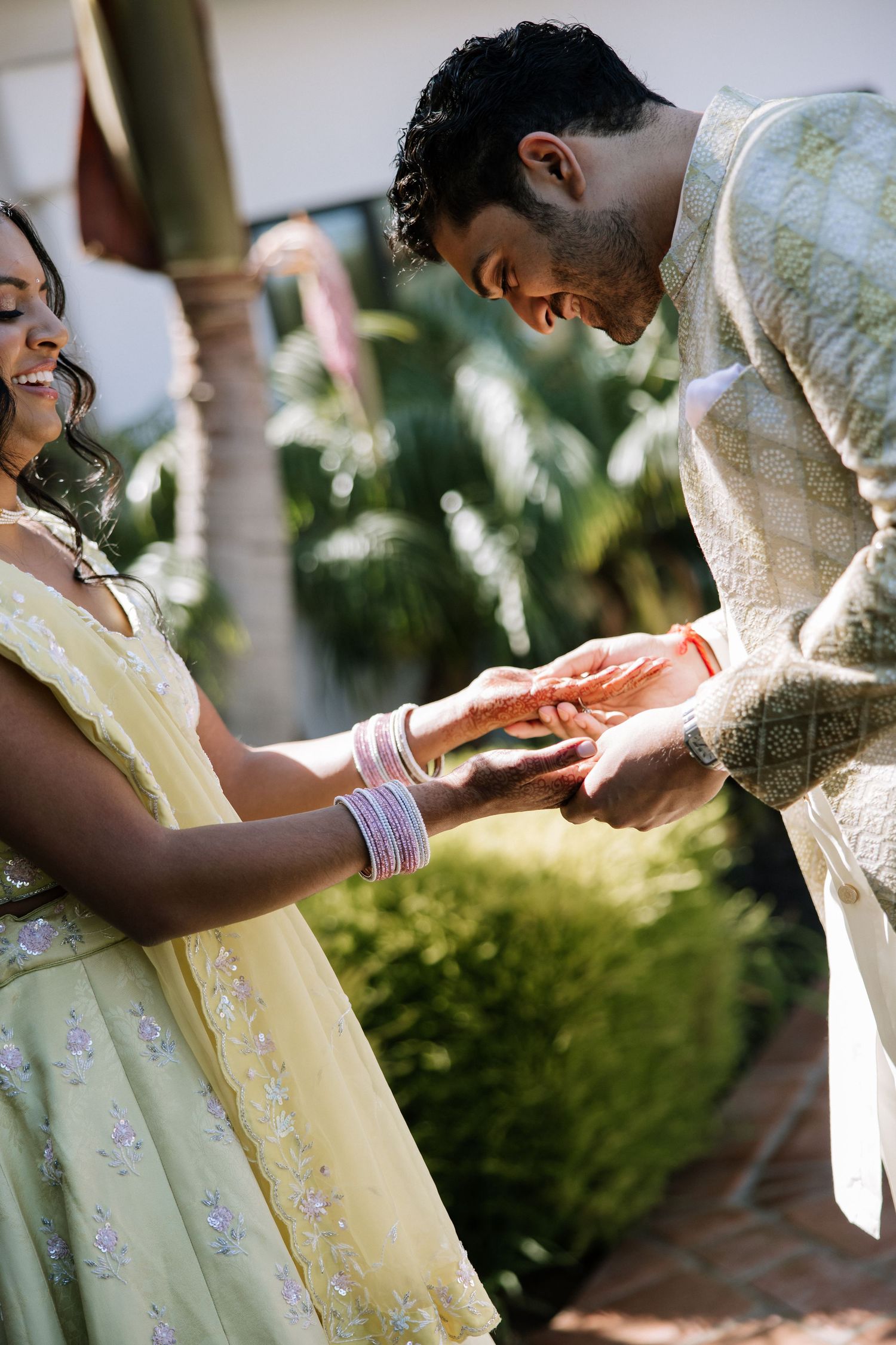 Smiling groom in embroidered sherwani places ring on bride's hand during traditional Indian wedding ceremony outdoors.