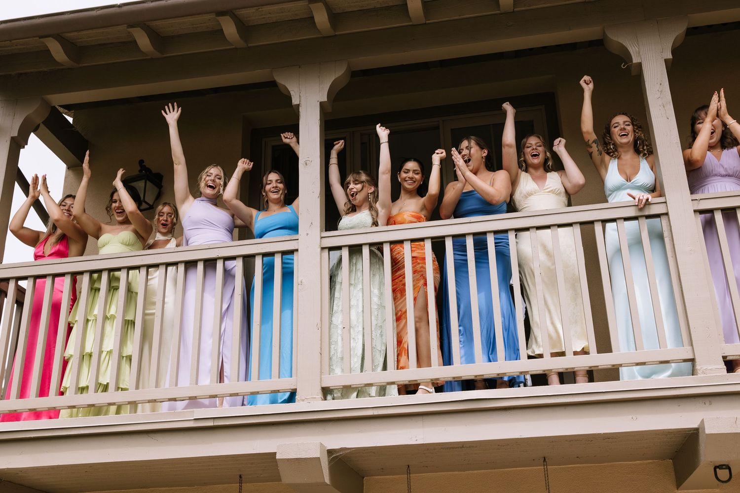 Bridesmaids in colorful dresses cheering with raised arms on a balcony.