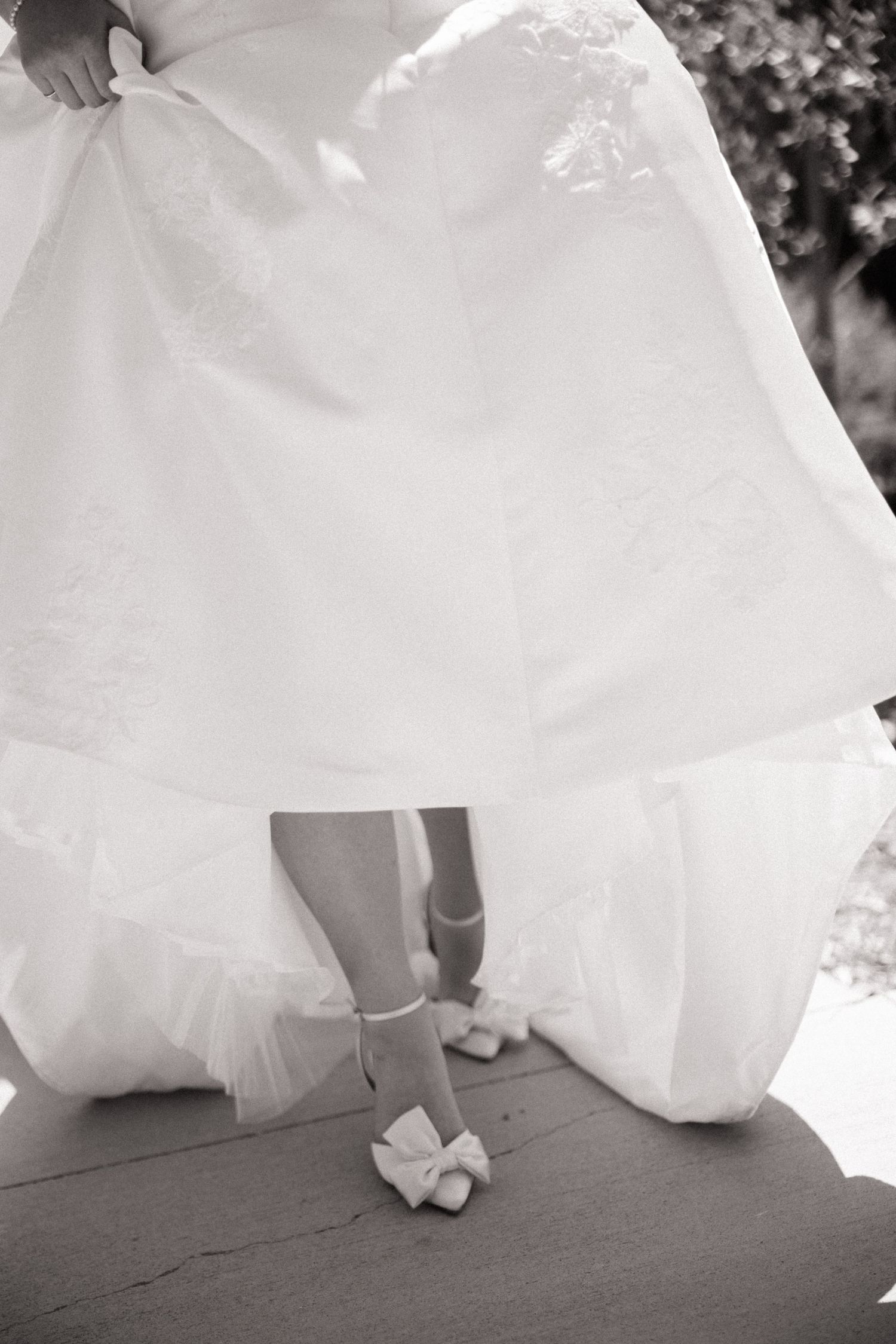 Black and white close-up of bride lifting wedding gown to reveal white bow heels on steps.