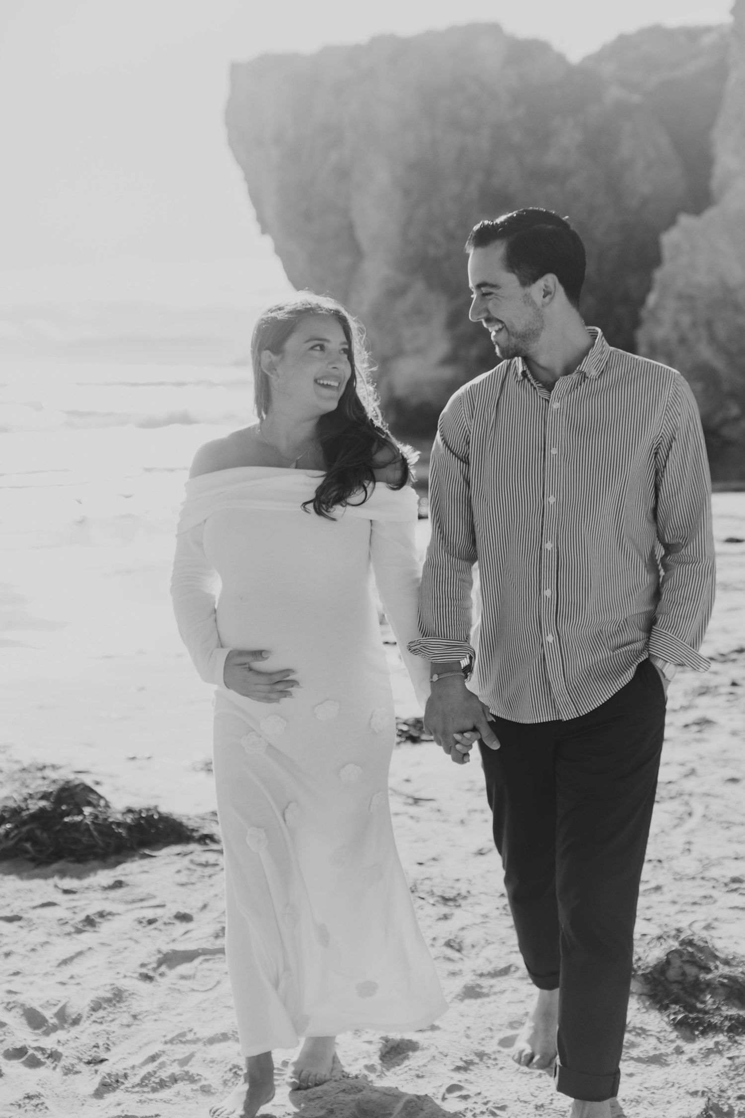 Black and white photo of a smiling pregnant couple holding hands and walking on a sandy beach near rocky cliffs.
