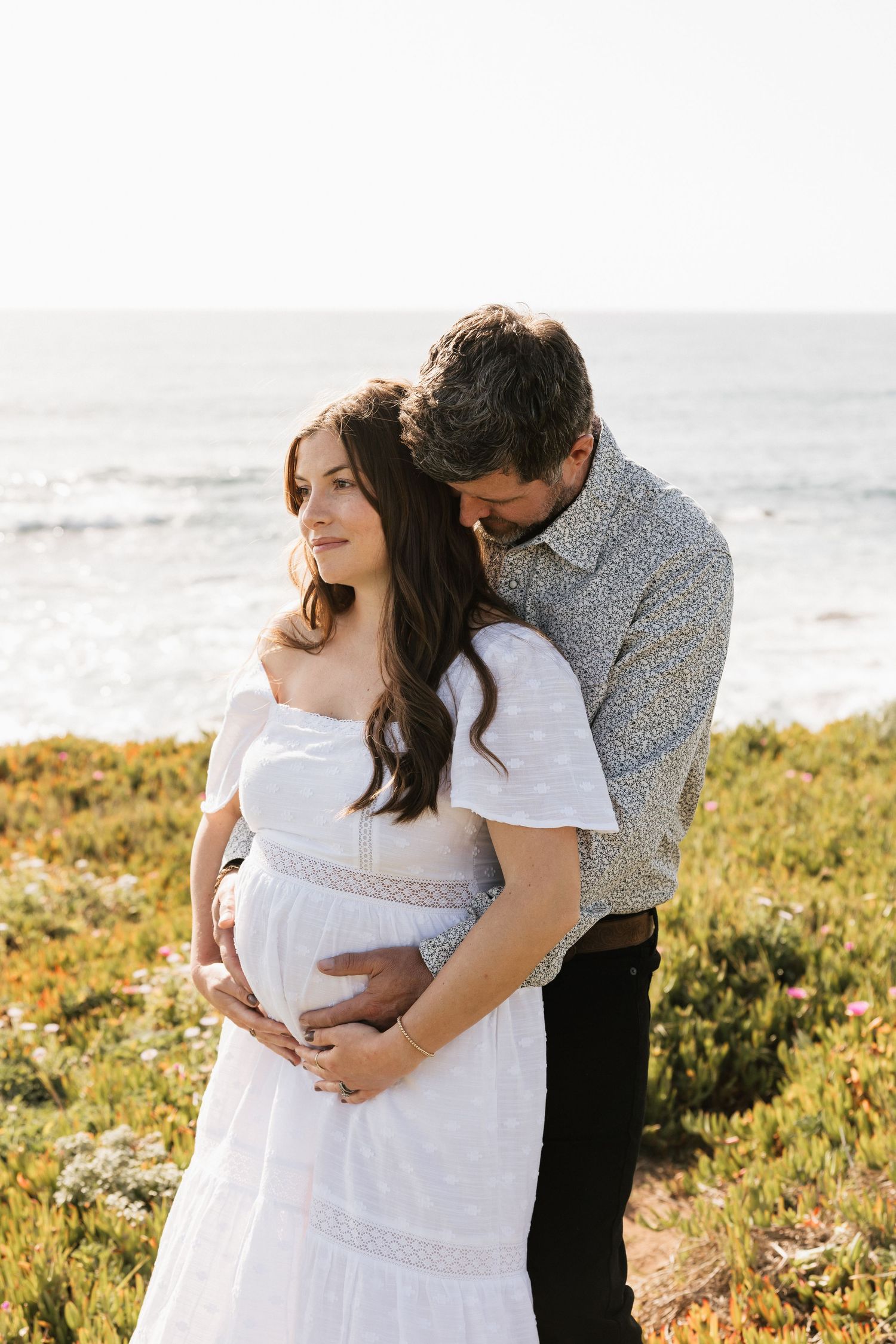 Man tenderly embracing pregnant woman in white dress from behind in colorful coastal flower field.