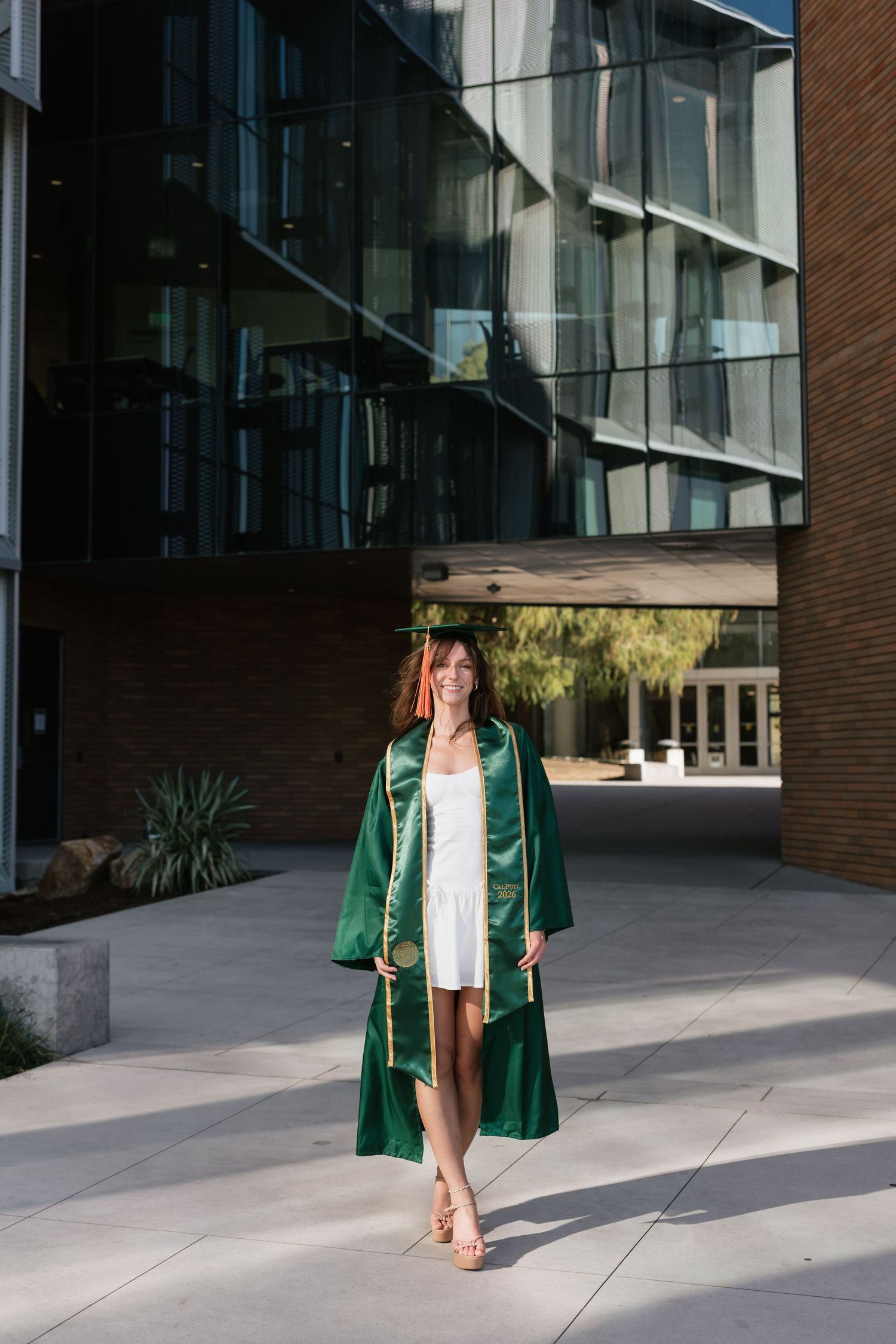 Smiling young woman in green graduation gown and cap standing outside modern university building.