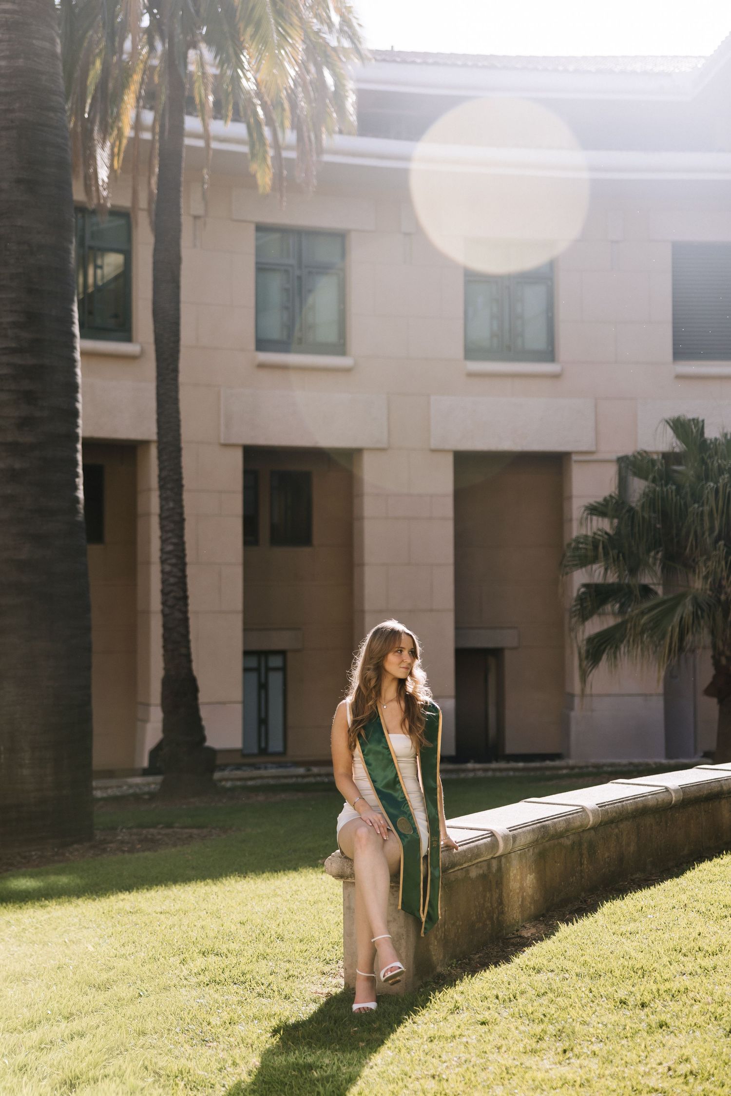 Graduate in white dress with green sash stands by palm trees in sunny campus courtyard.