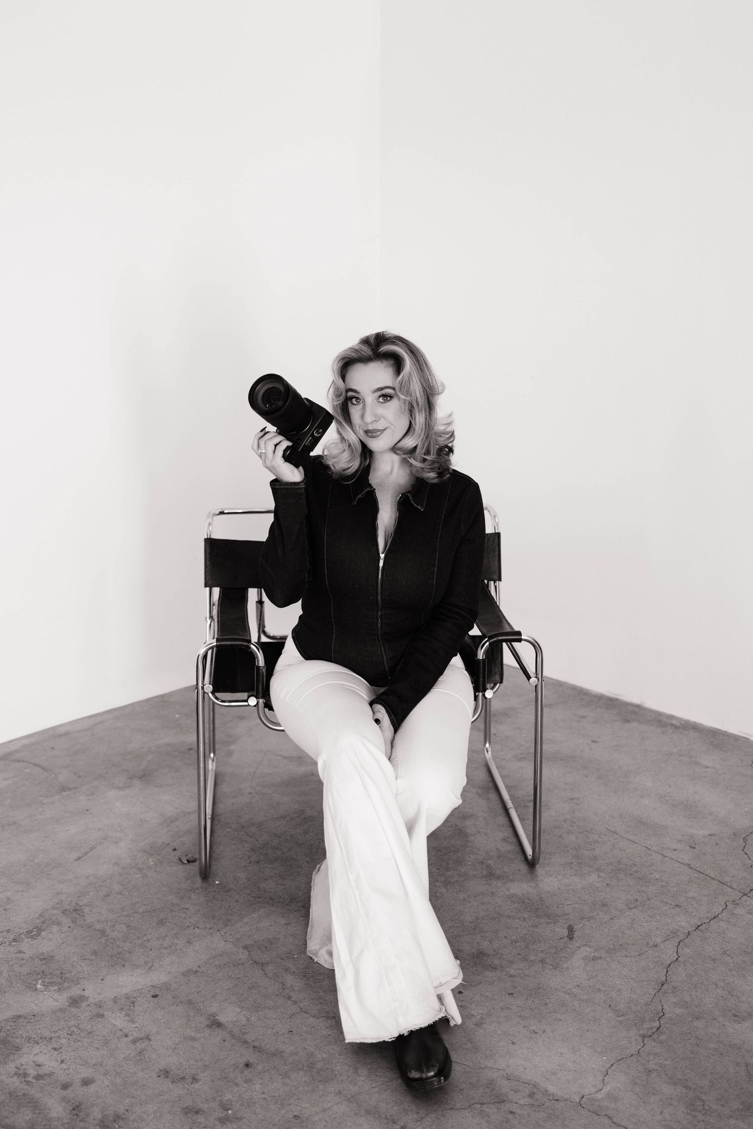 Blonde woman in black jacket and white flares holding camera, seated in chair, black and white photo.