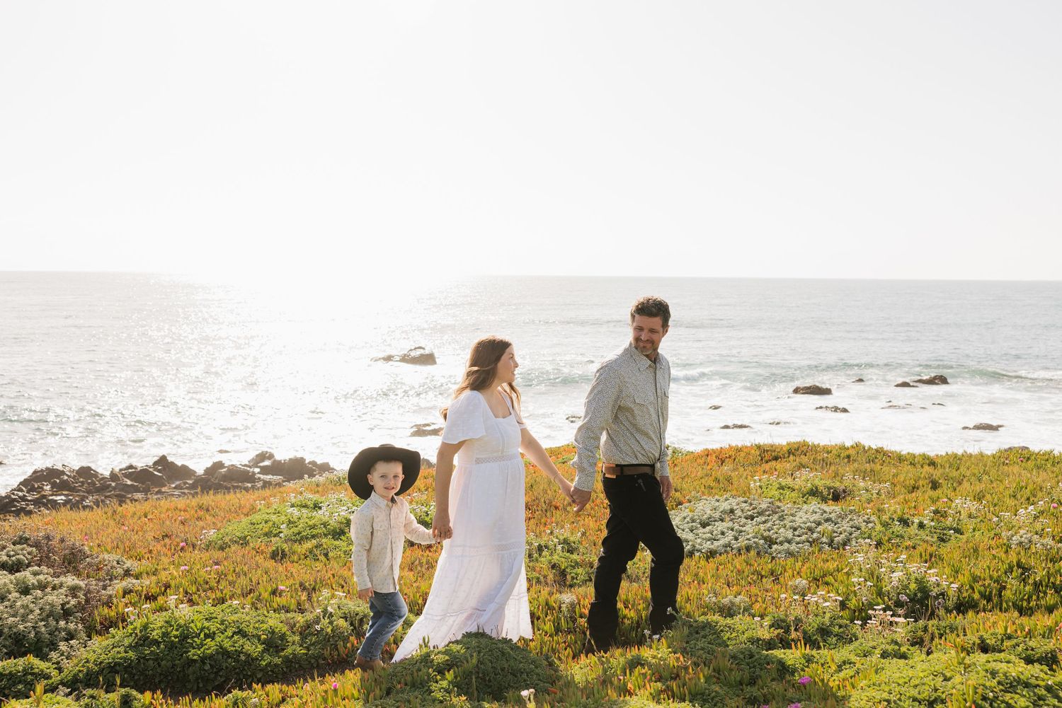 Family maternity photo with couple and young child walking through coastal wildflower field at sunset.