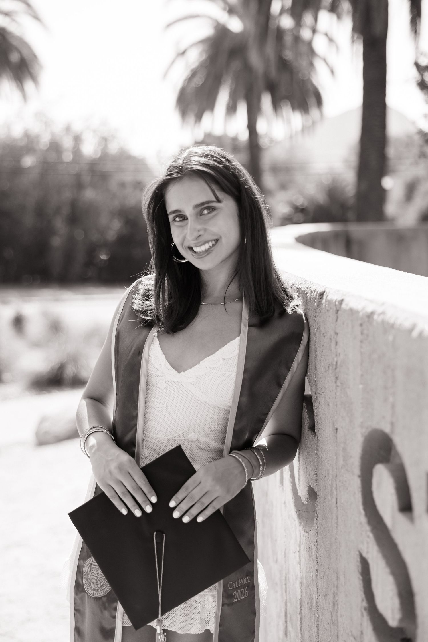 Smiling young woman in graduation gown holding cap outdoors near palm trees in black and white photo.