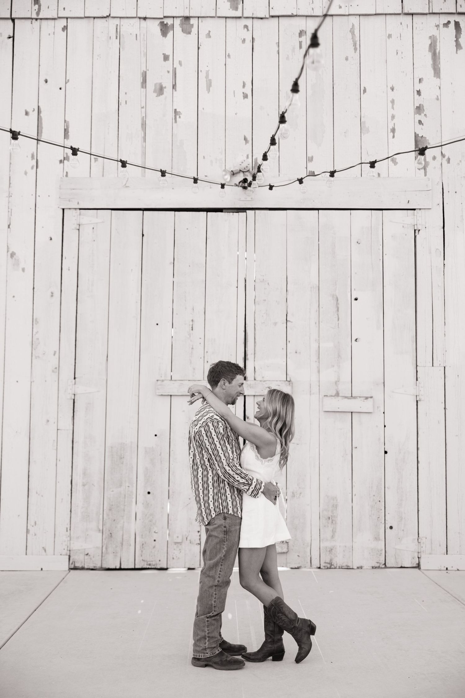 Couple sharing romantic kiss in front of rustic white barn doors with string lights above.