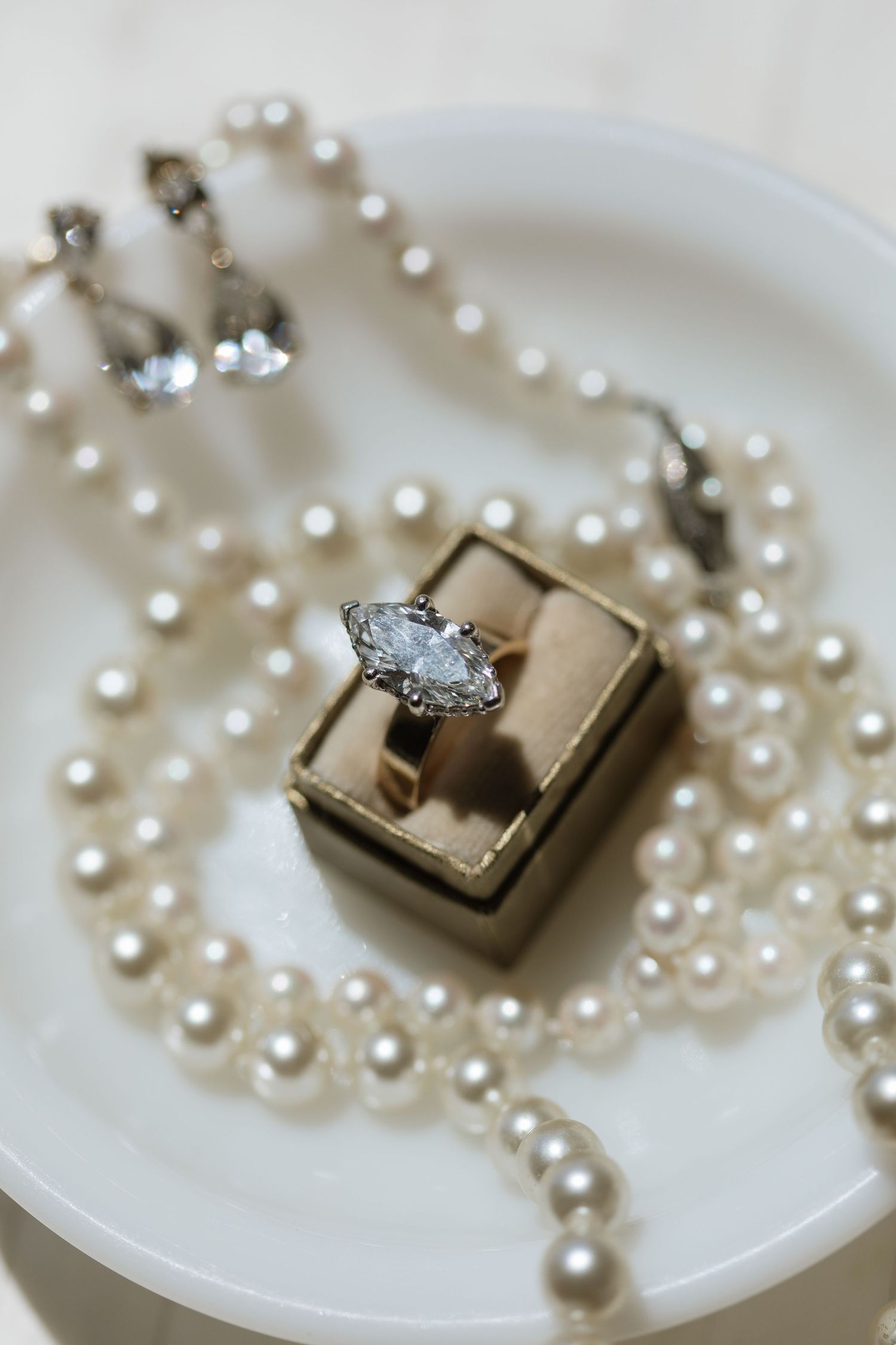 Marquise diamond engagement ring in gold box surrounded by pearl necklace and earrings on white plate.