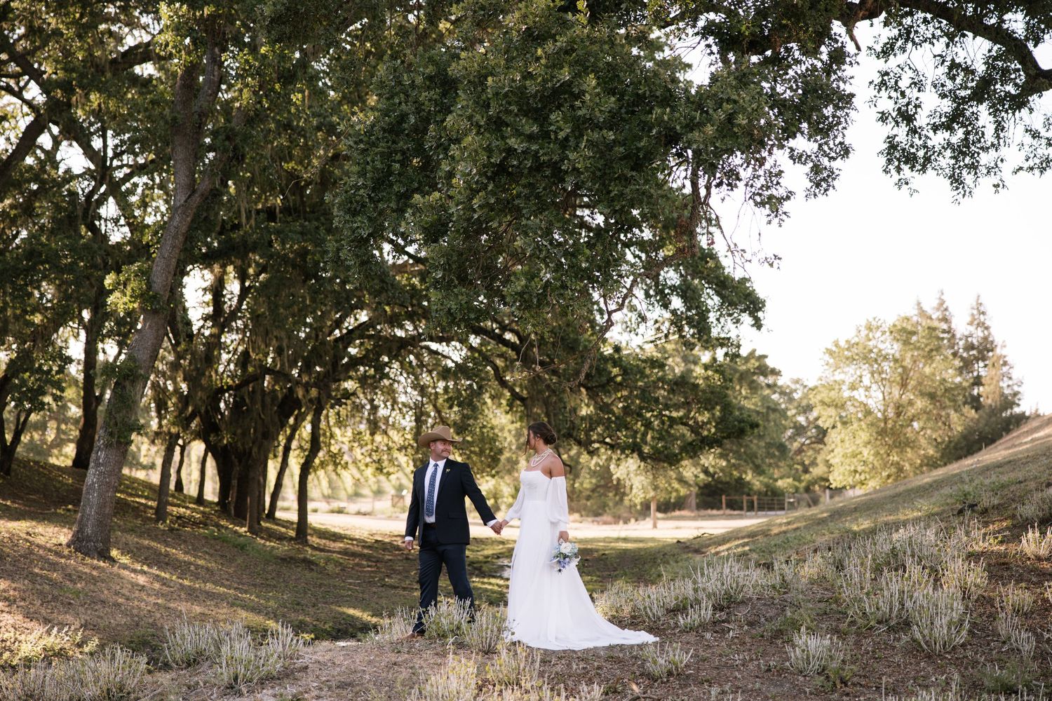 Bride and groom walk hand in hand through golden sunlit oak trees in rustic outdoor wedding setting.