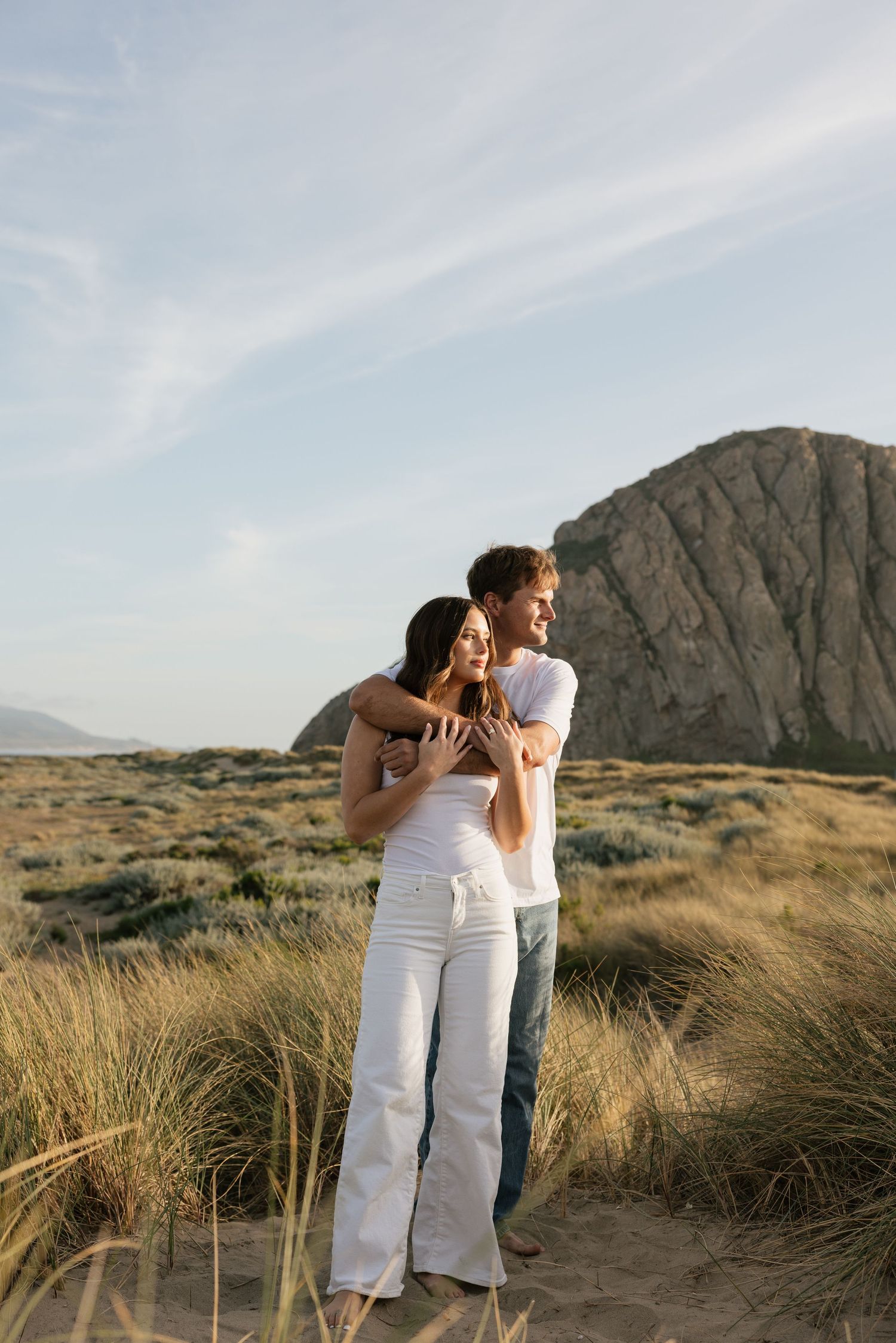 Romantic couple hugging in coastal dunes with large rock formation behind them.
