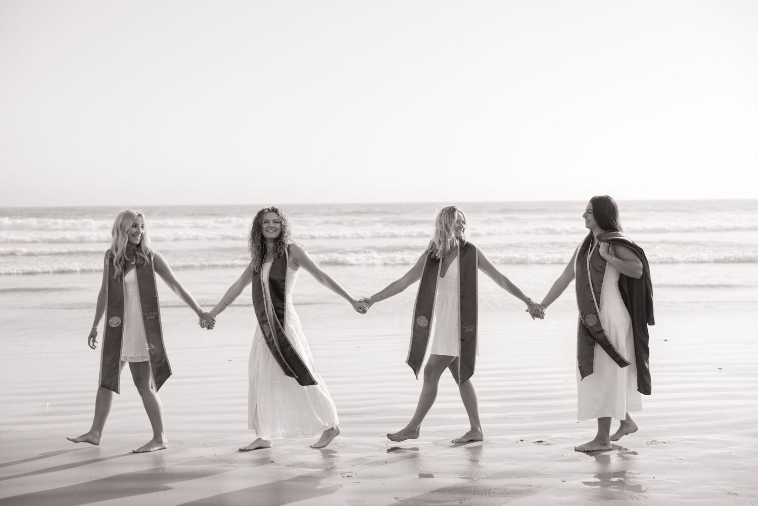 Four female graduates hold hands walking along ocean beach in black and white graduation photo.
