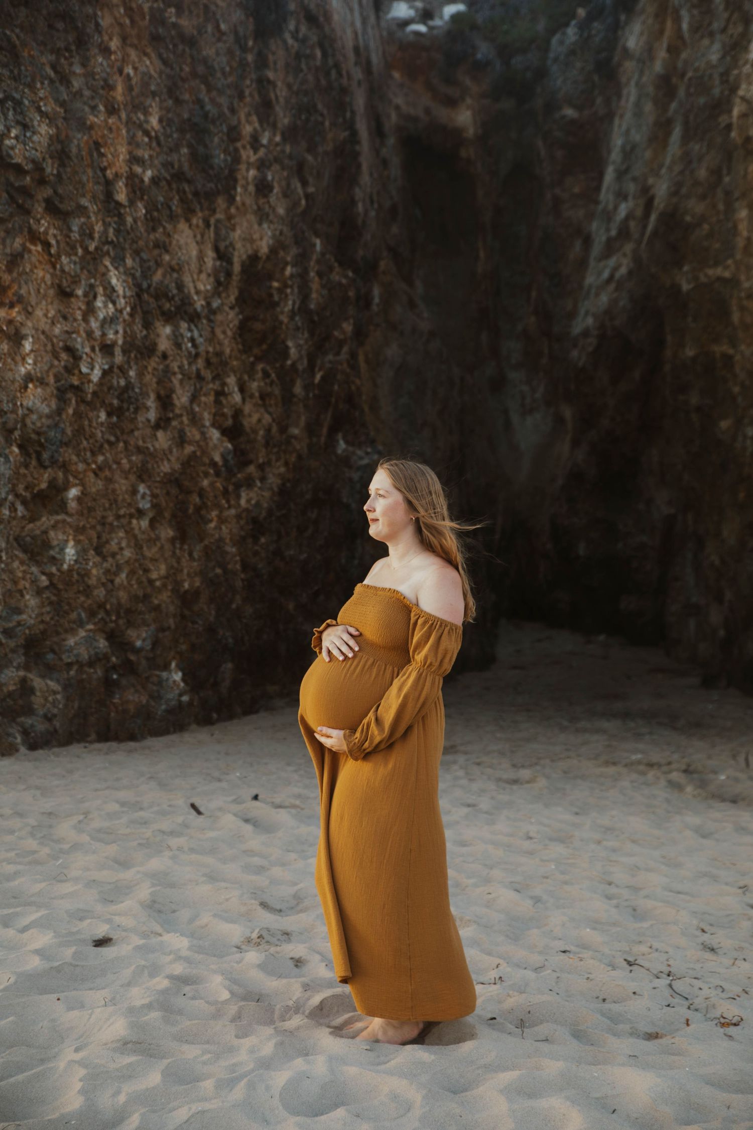 Pregnant woman in golden off-shoulder maxi dress standing on sandy beach near rock cliffs.