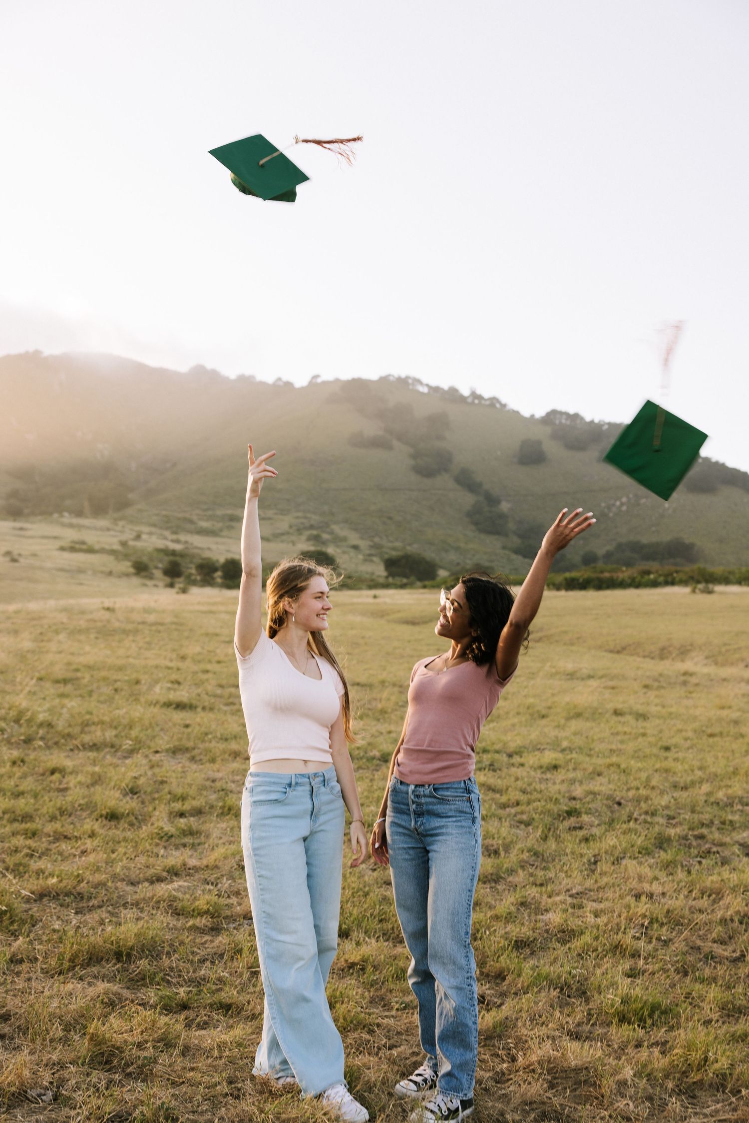 Two women tossing green Cal Poly graduation caps in the air in a sunny green meadow.