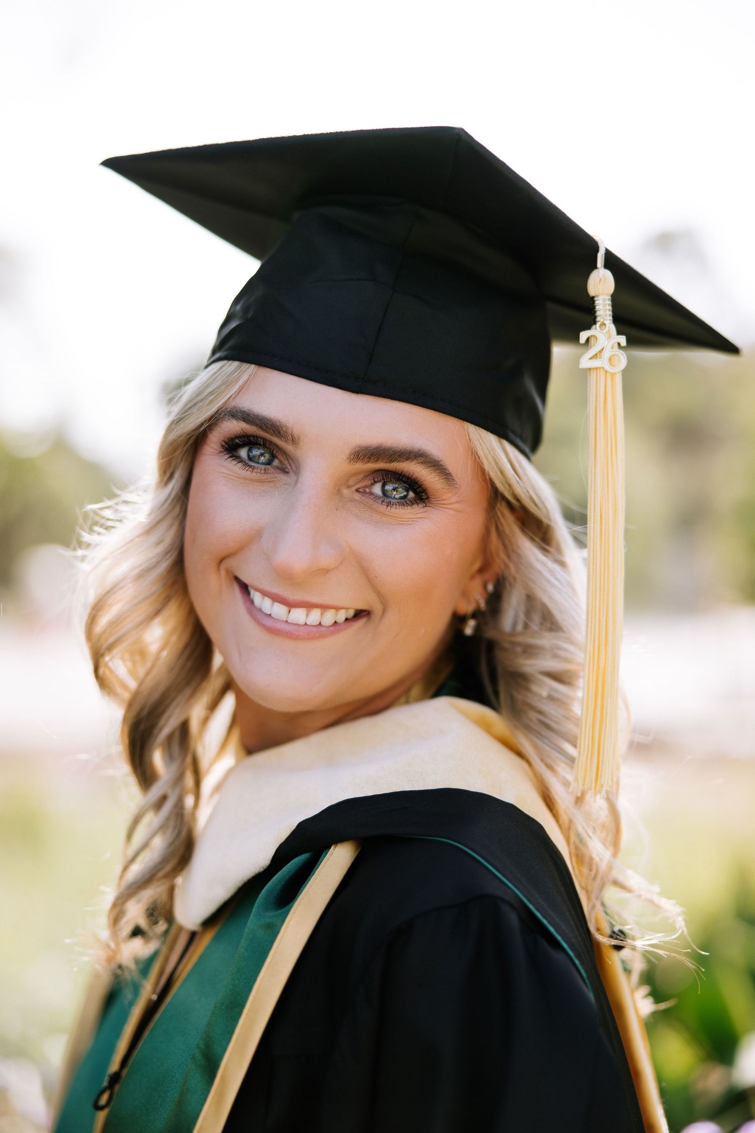 Close-up portrait of a smiling blonde woman in a black and green graduation cap and gown with gold tassel.