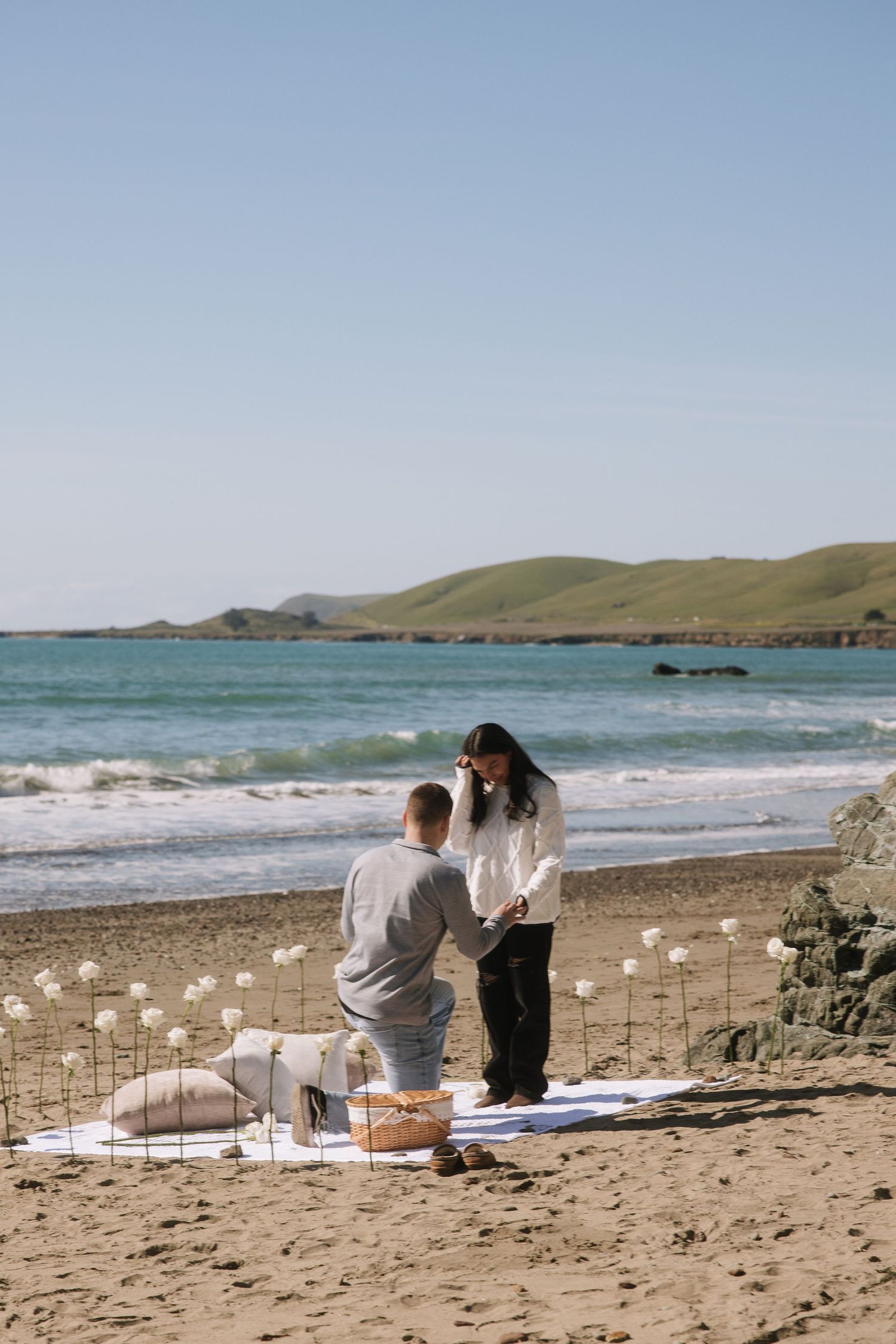 Man proposing to his girlfriend on a scenic beach with white roses and a picnic setup.