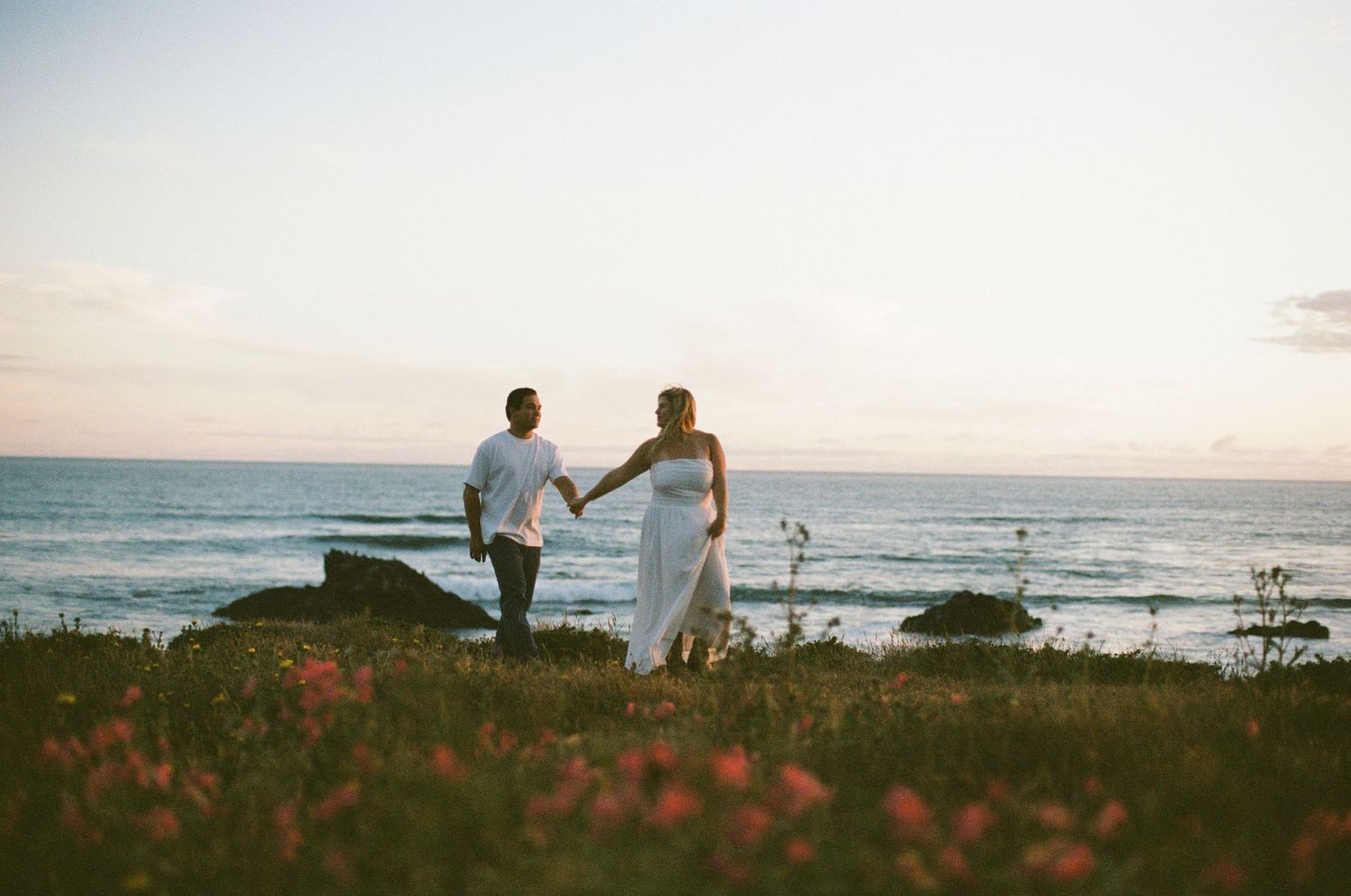 Engaged couple holds hands walking near ocean at sunset with pink wildflowers.