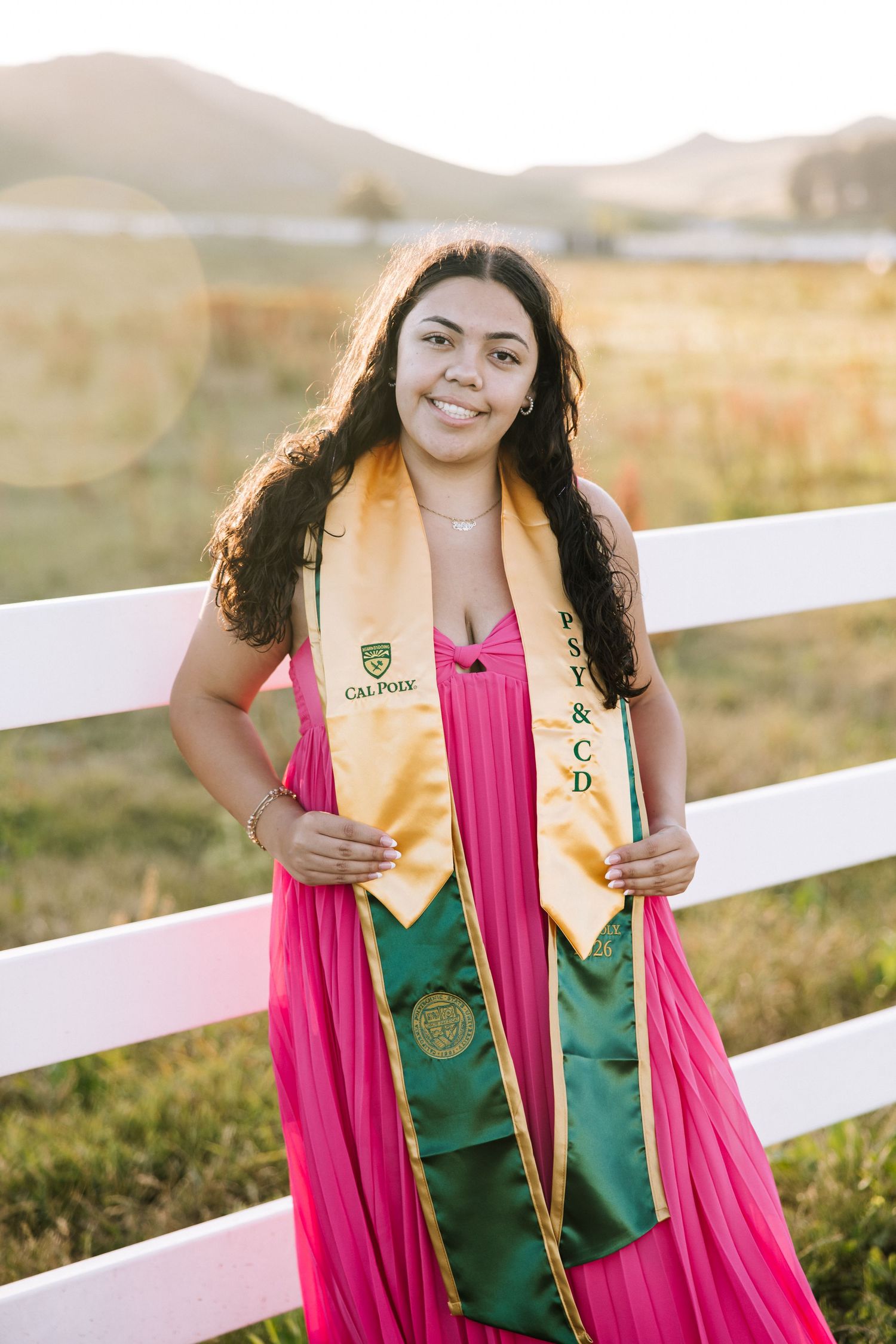 Smiling woman in pink dress wearing Cal Poly green and gold graduation sashes leaning on white fence.