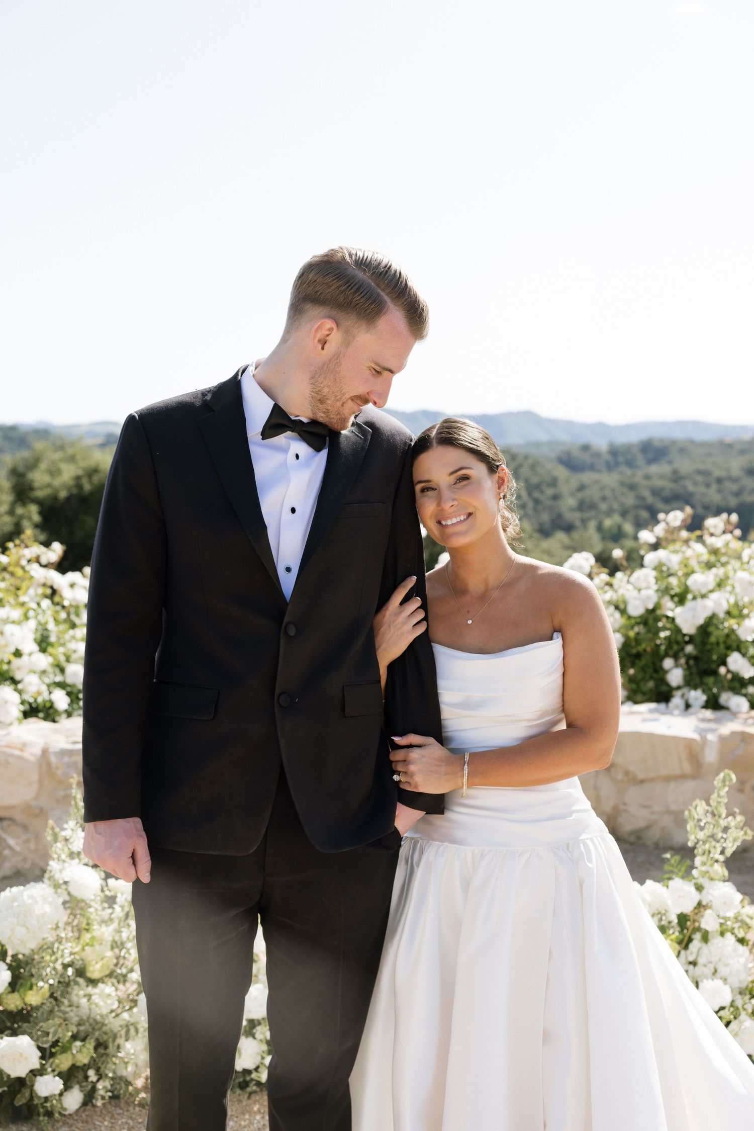 Bride and groom pose outdoors with white floral arrangements and rolling hills in the background on their wedding day.