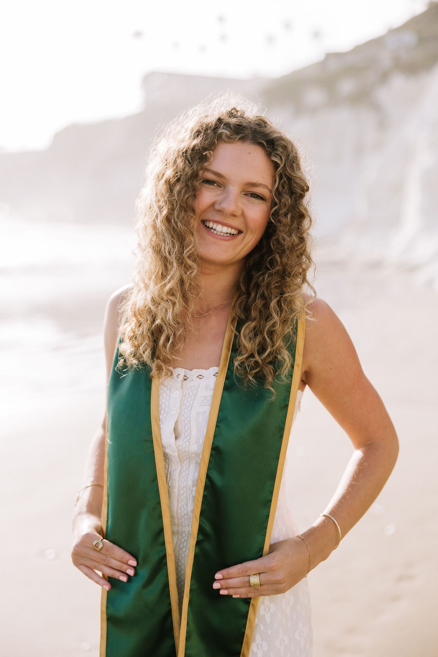 Smiling female graduate with curly hair wearing green and gold graduation stole on sunny beach.