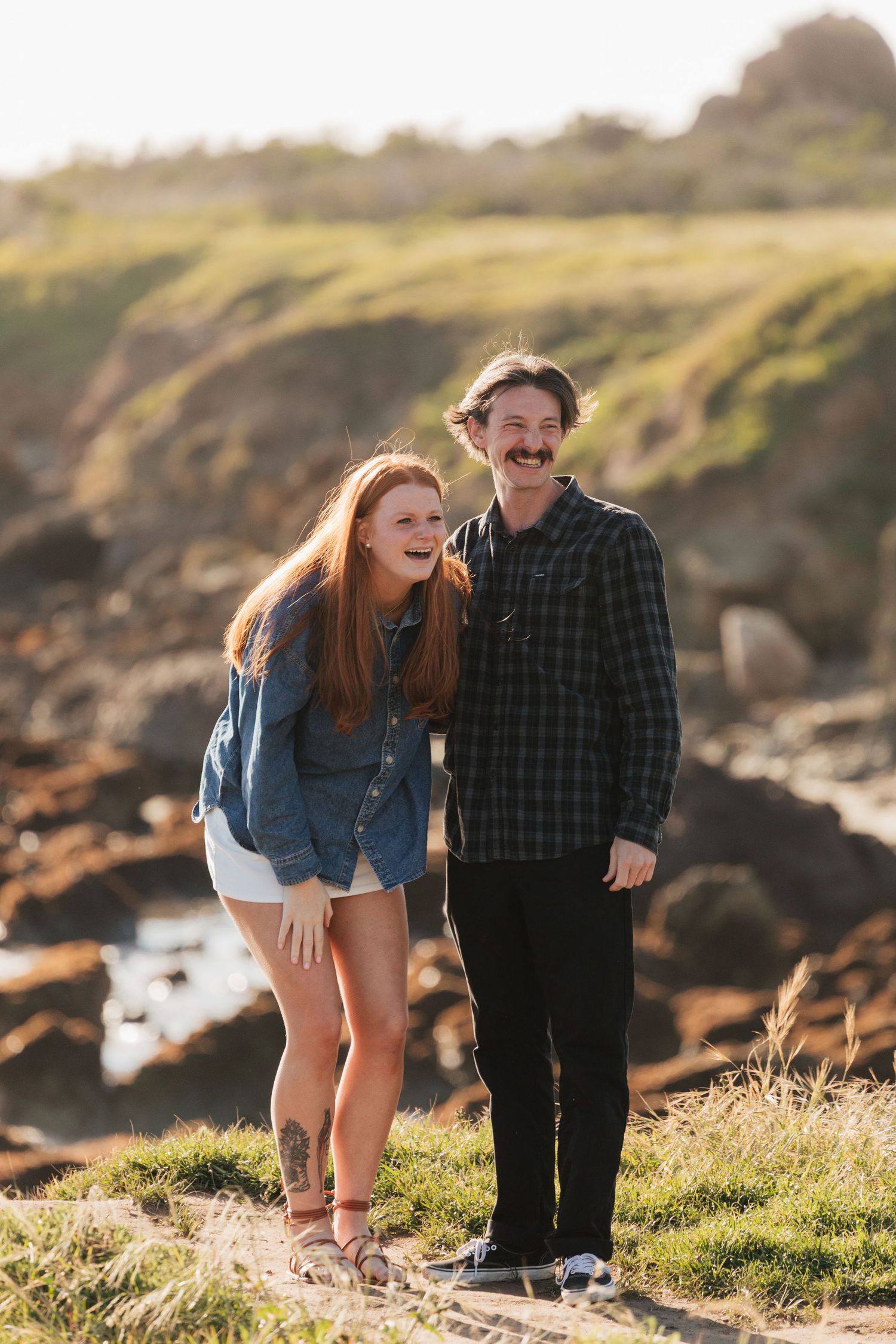 Happy couple laughing together on rocky coastal cliffs during golden sunset engagement photo session.