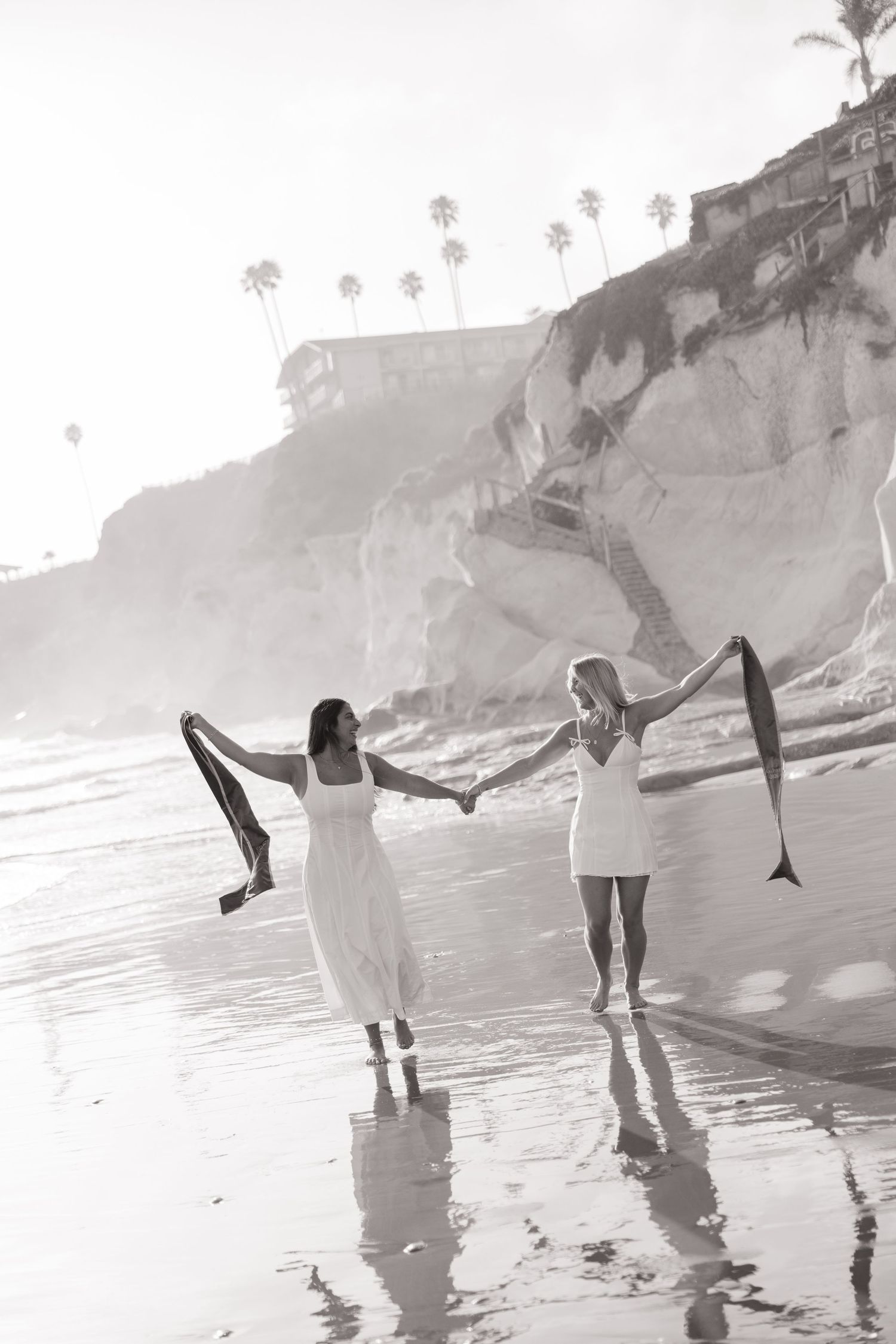Black and white photo of two women in white dresses walking on a beach holding a sash.