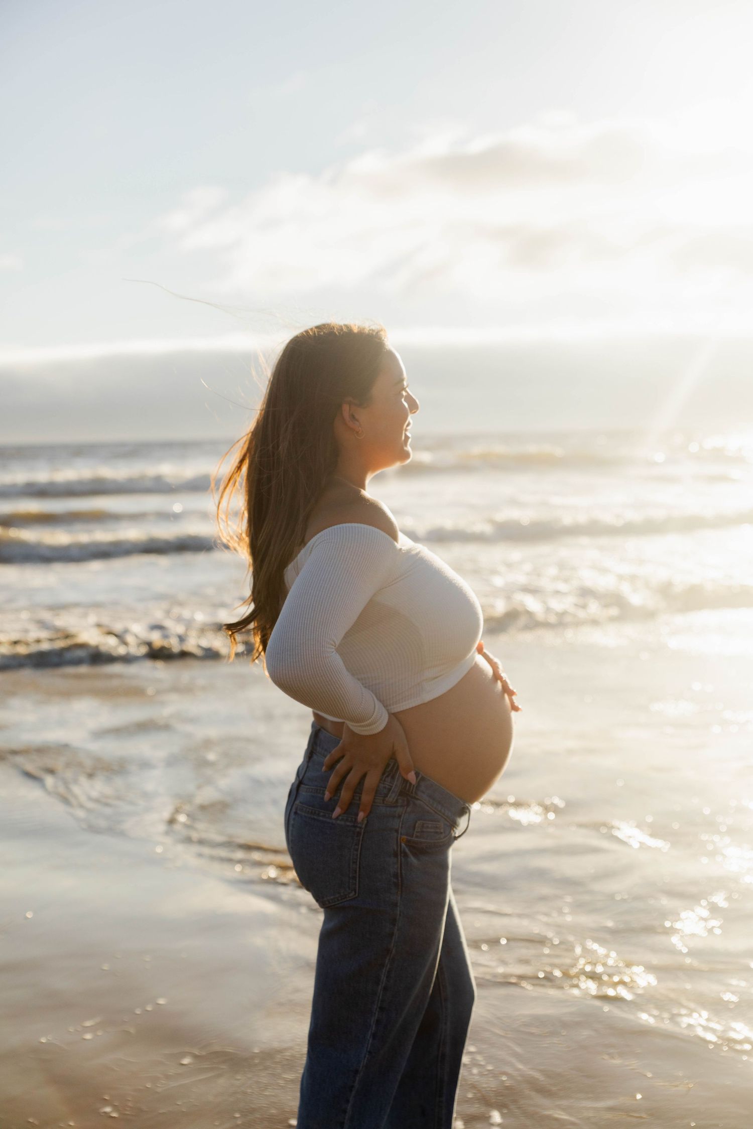 Pregnant woman in white top and jeans standing on a sunny beach, cradling her baby bump with ocean waves behind her.