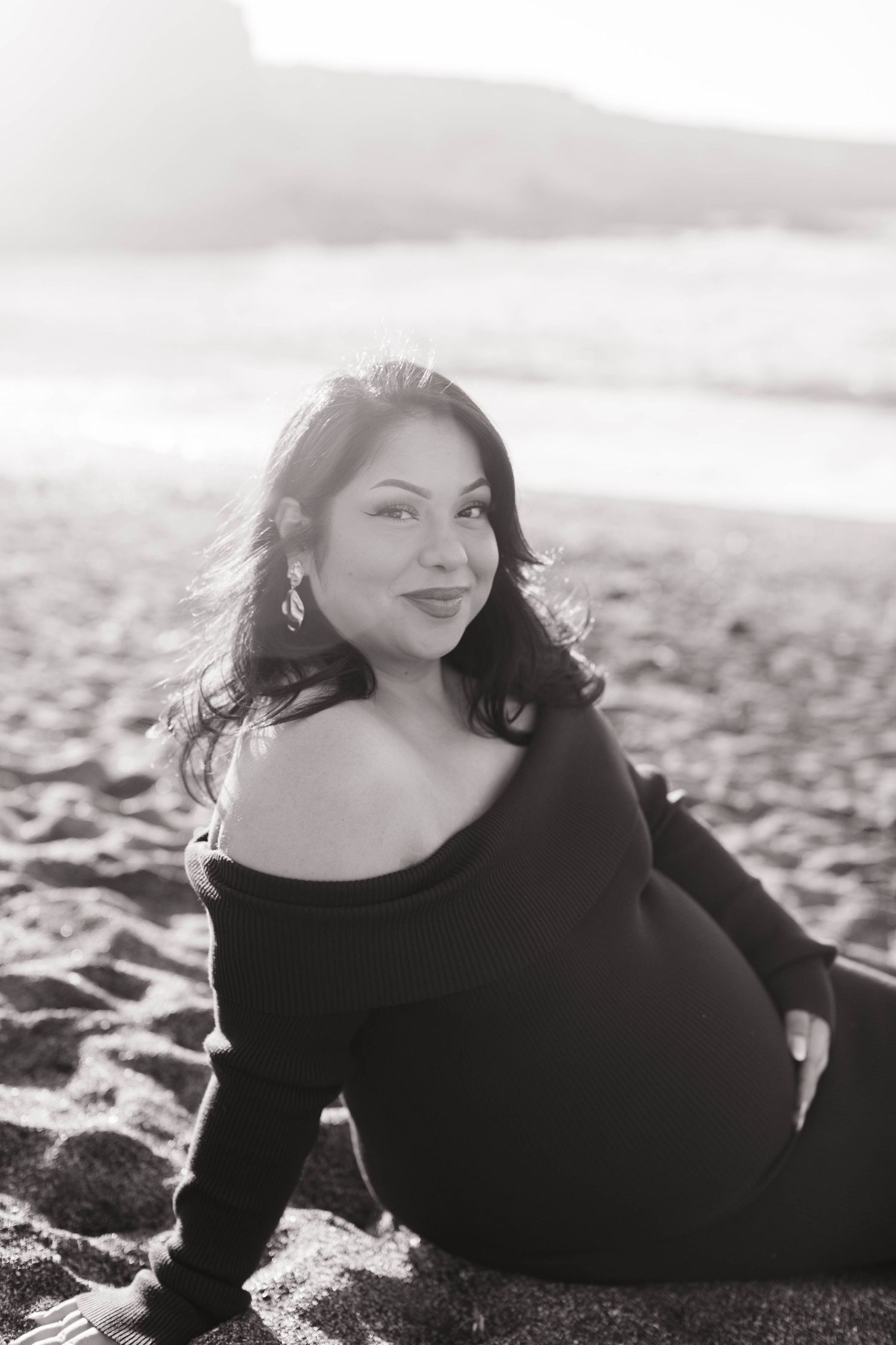 Black and white beach maternity portrait of smiling pregnant woman in black off-shoulder dress.