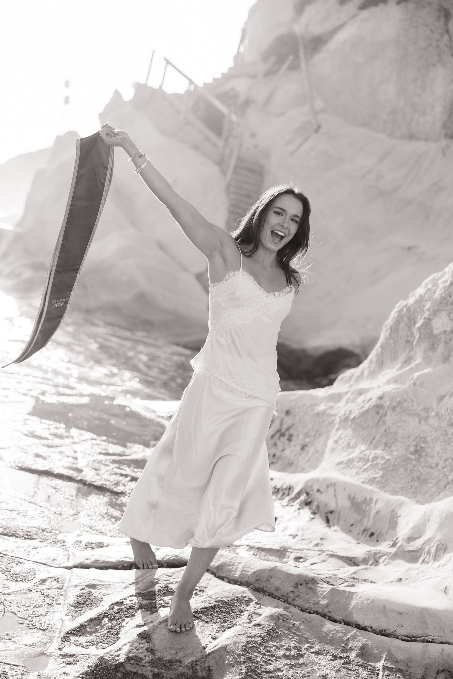 Black and white photo of laughing woman waving graduation stole on rocky coastal shore.
