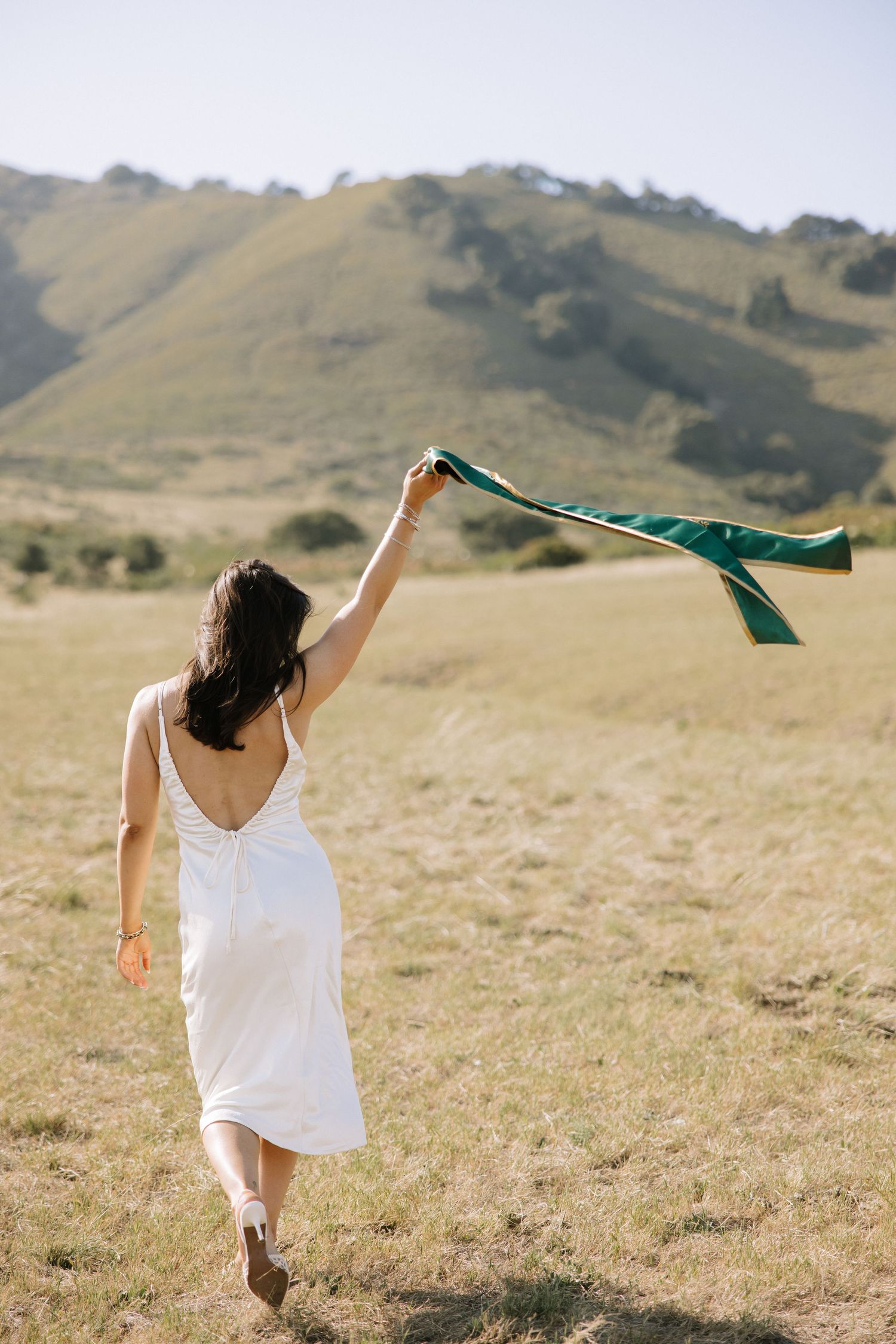 Young woman in white dress holding green graduation stole while walking through sunny green hillside.