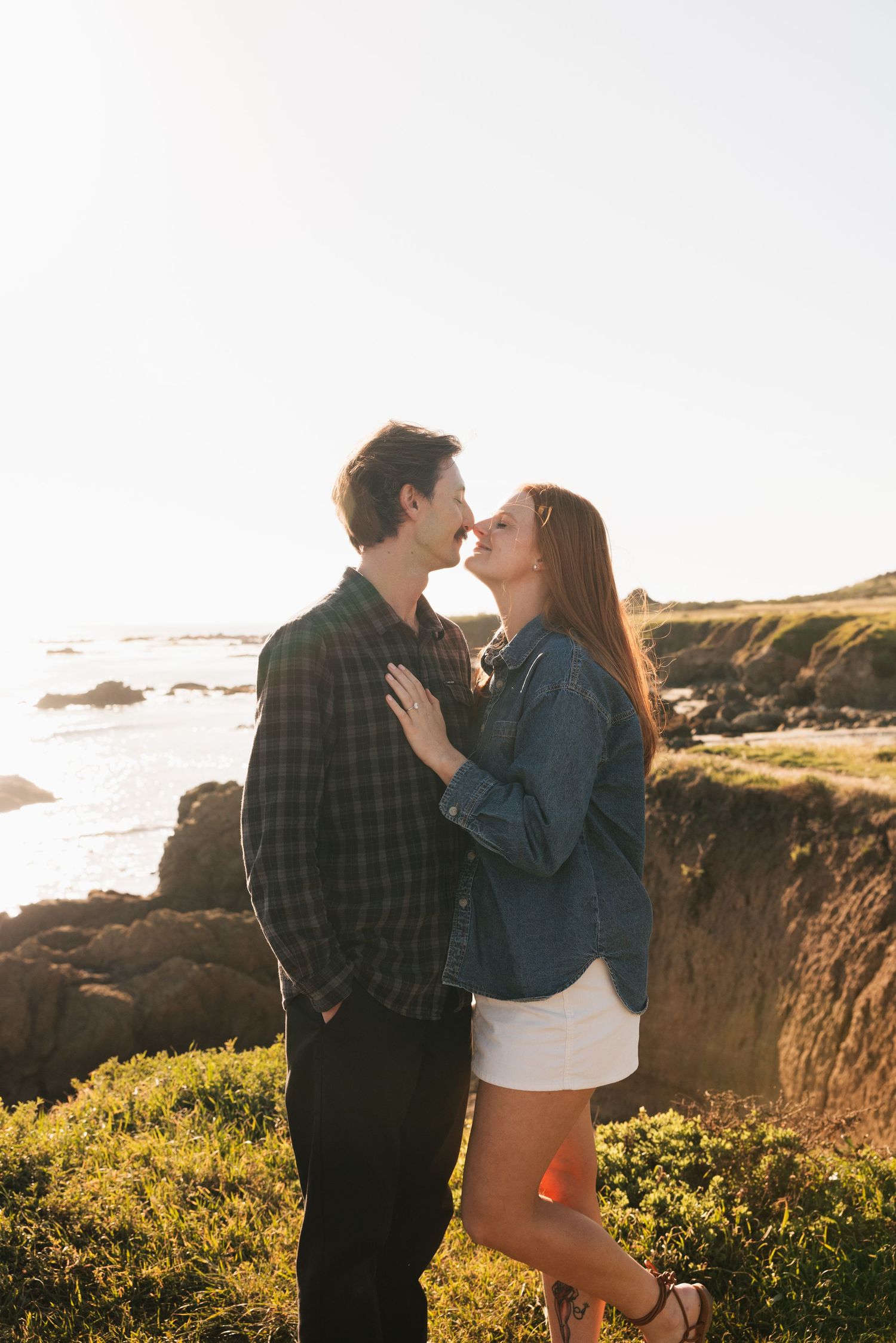 Couple sharing romantic almost-kiss moment on coastal cliffs with ocean view during golden hour.