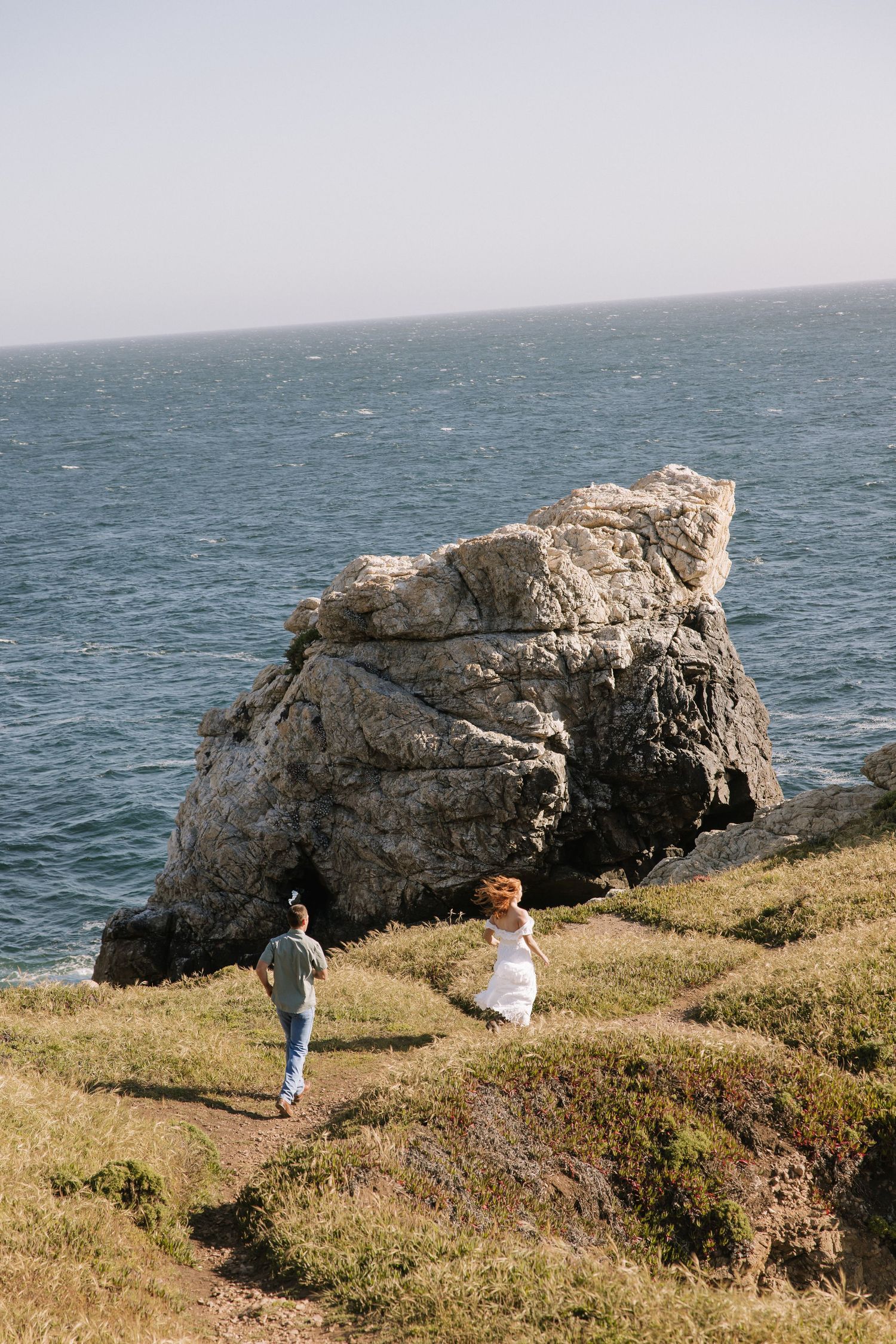 Couple walking along a coastal cliff path with a large rock formation and blue ocean in the background.