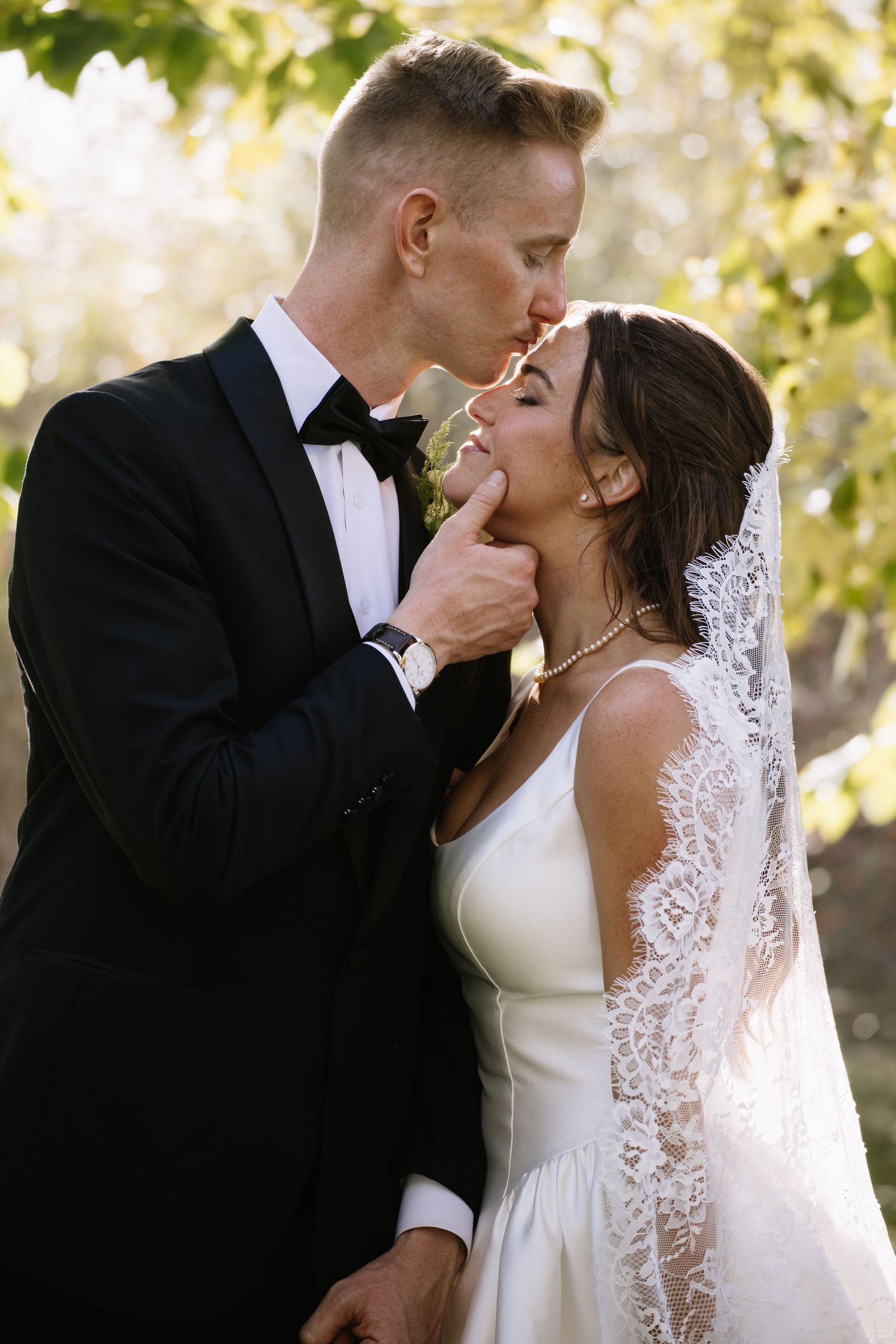 Groom in black tuxedo gently tilts bride's chin for a kiss outdoors, bride wearing lace veil and white gown.