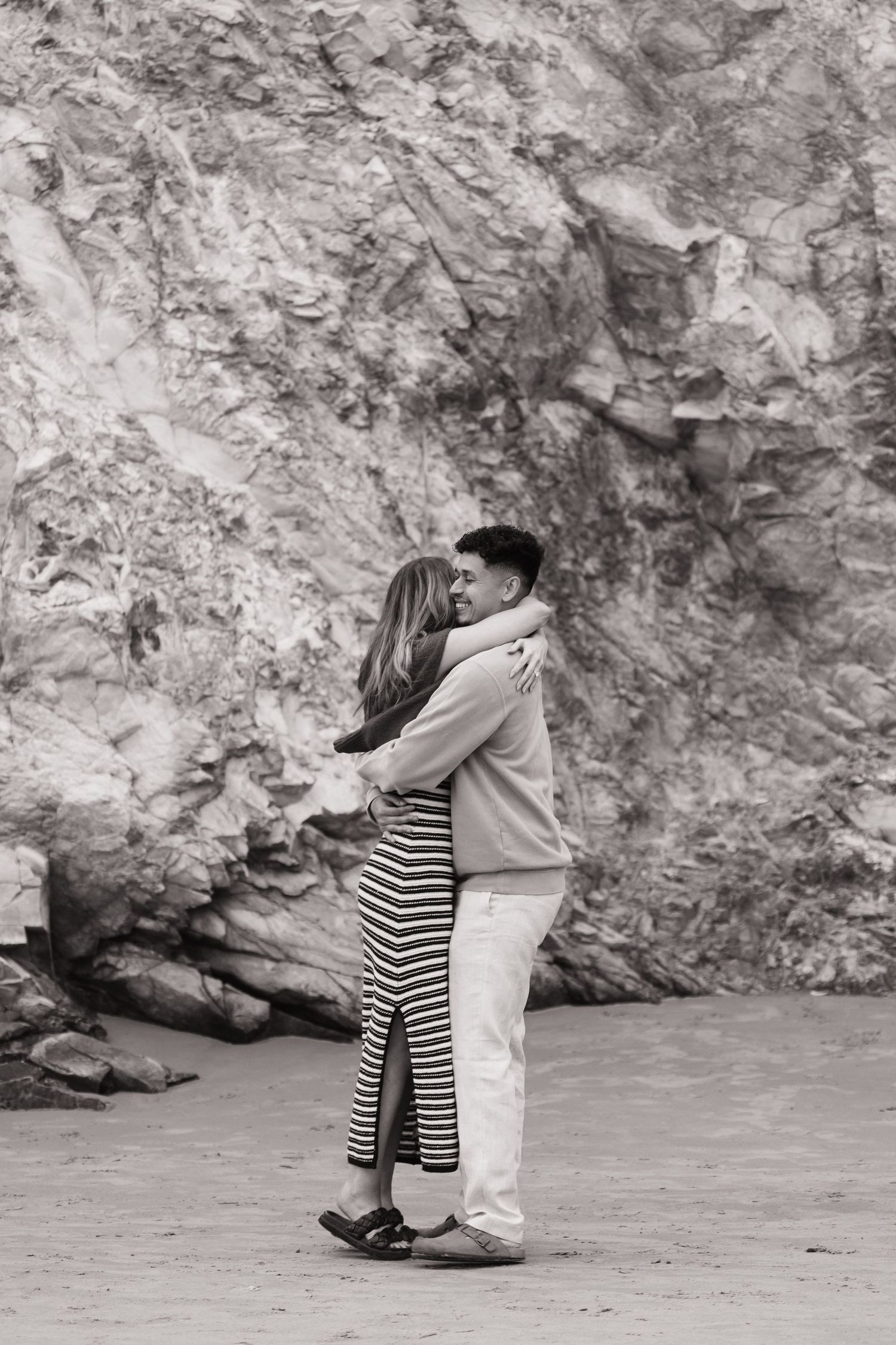 Black and white photo of a couple embracing on a beach with rocky cliffs in the background.