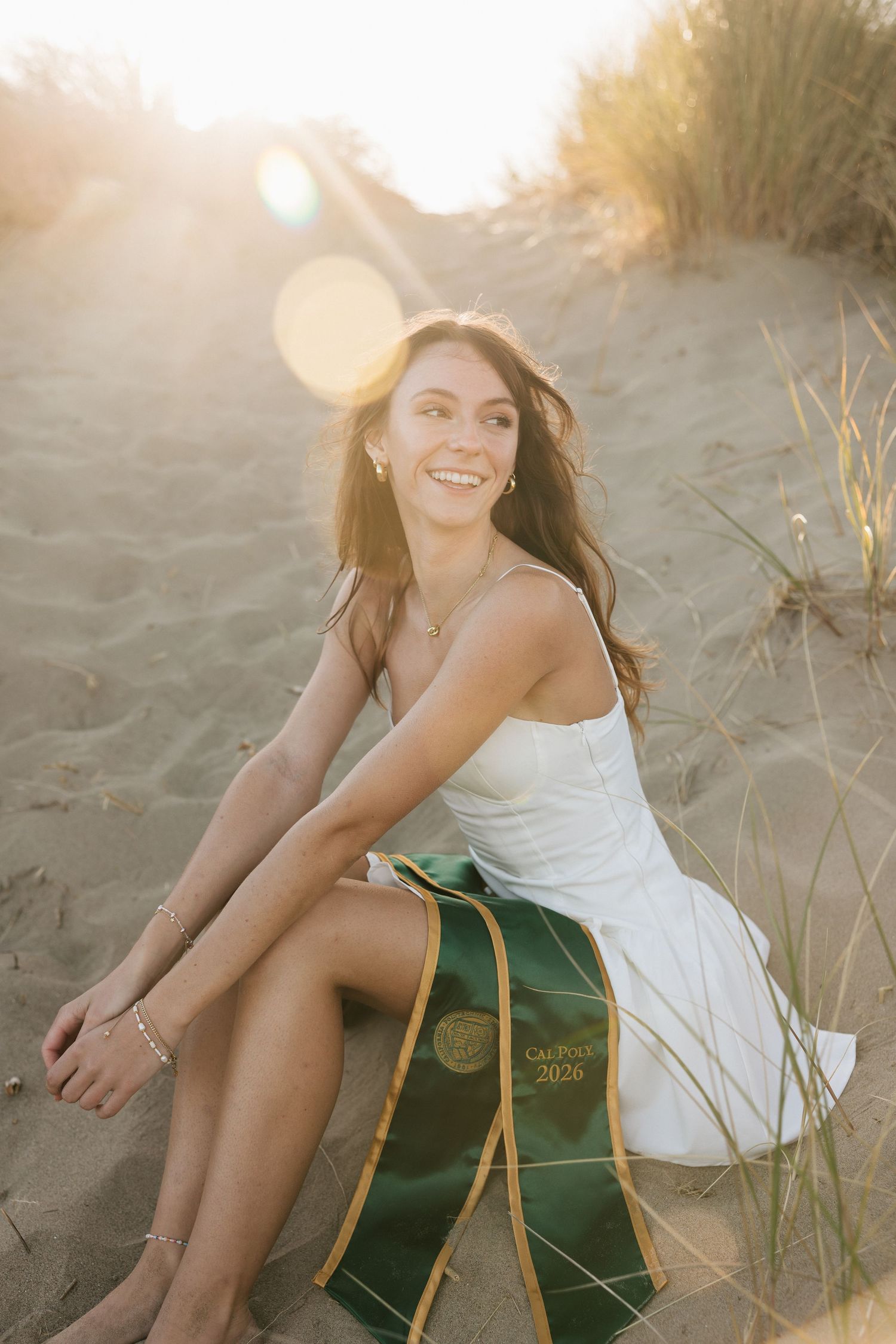 Smiling woman in white dress sitting on beach sand dunes holding Cal Poly 2026 graduation sash at sunset.