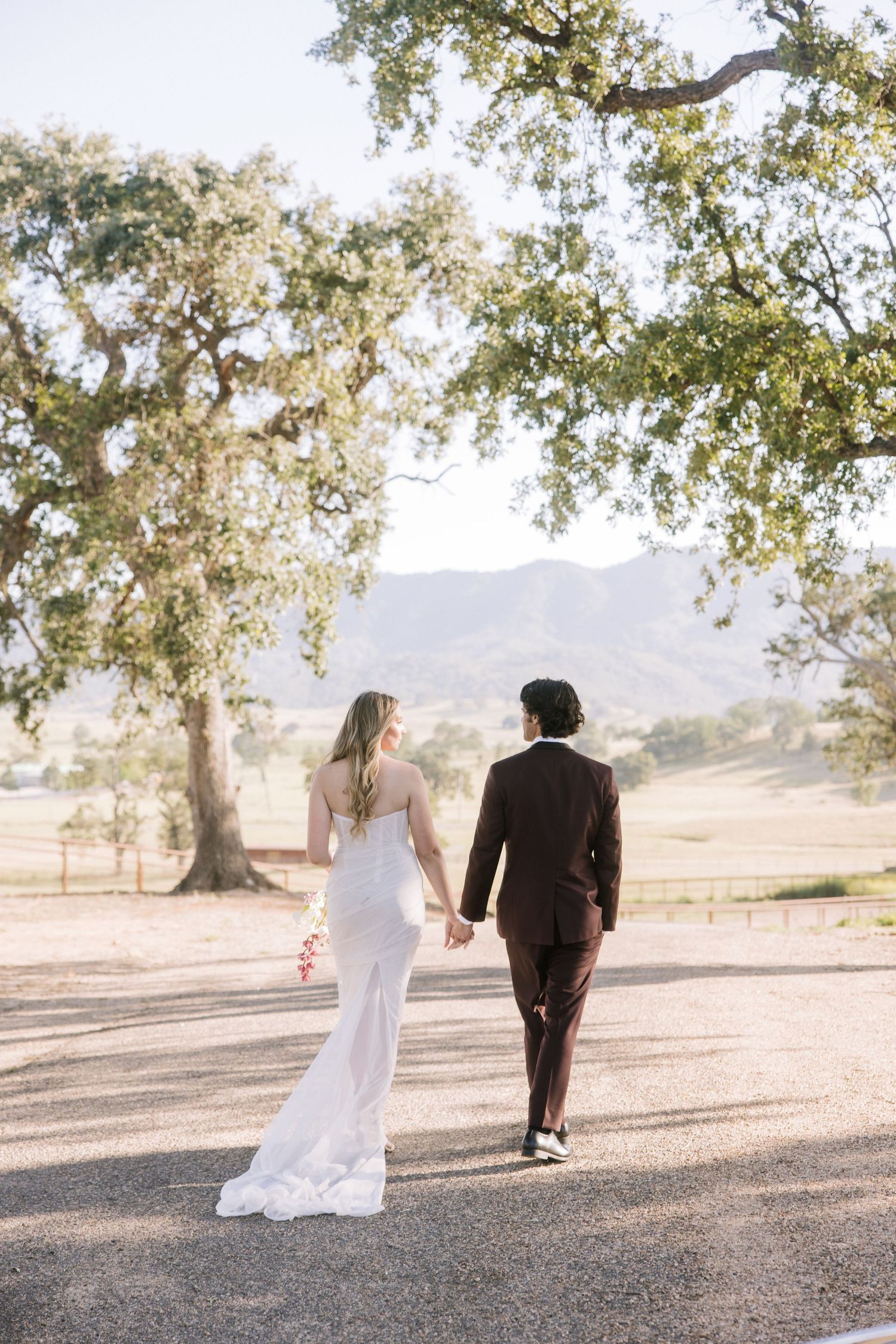 A bride and groom walk hand-in-hand down a sunlit path framed by oak trees and mountain views.