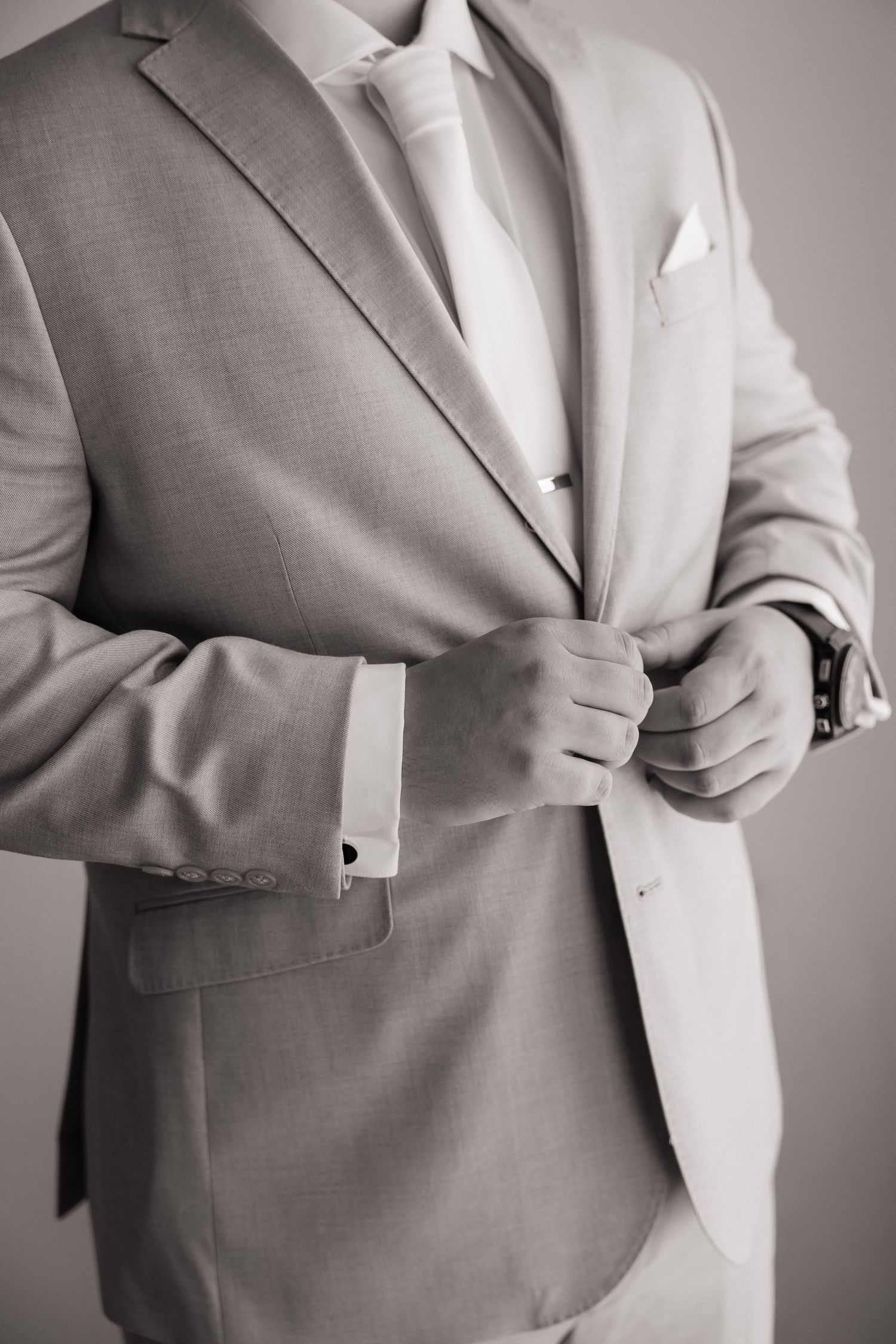 Black and white close-up of groom buttoning light gray suit jacket with white tie and pocket square.