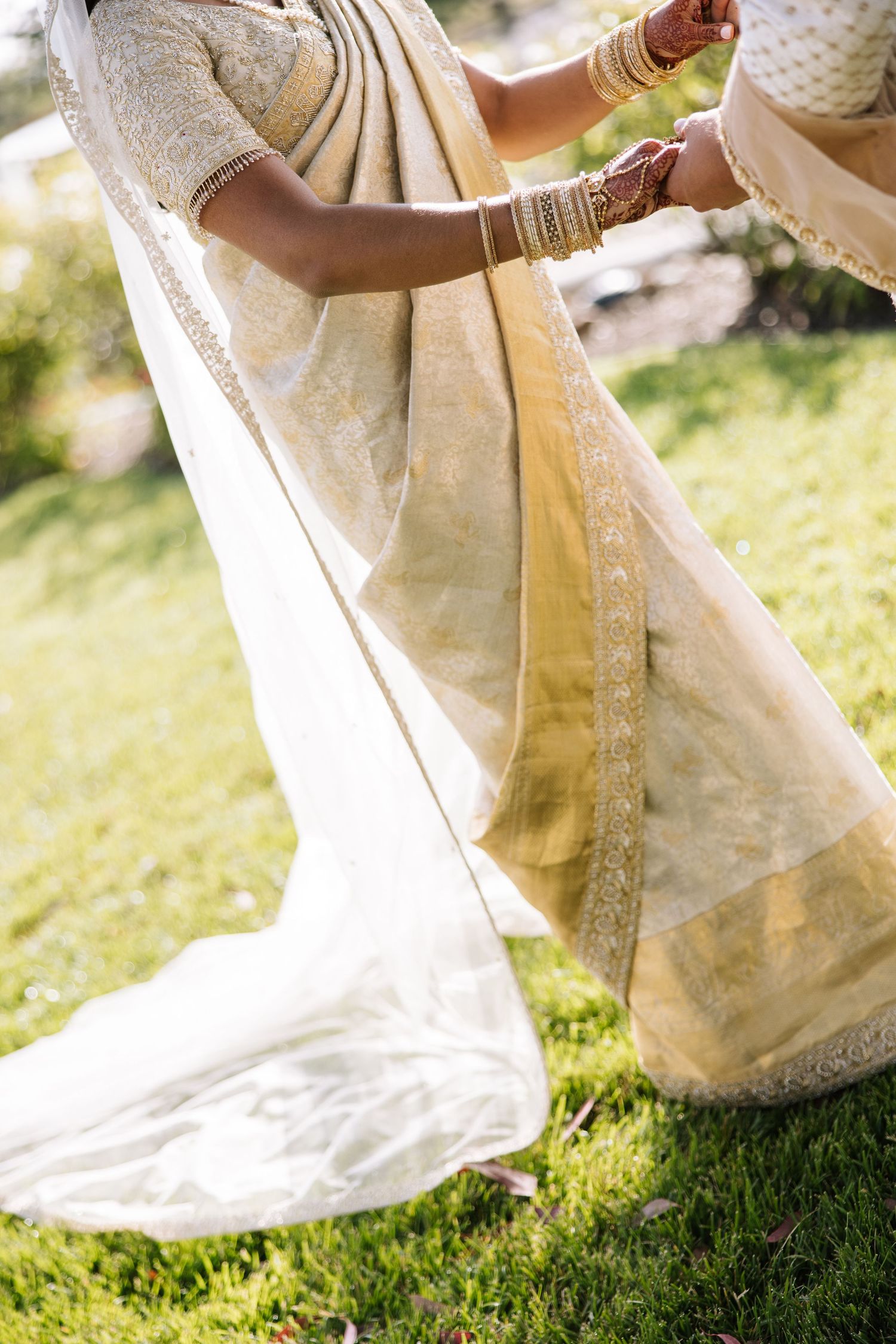 Close-up of bride in ivory and gold embroidered saree being dressed outdoors on green grass.