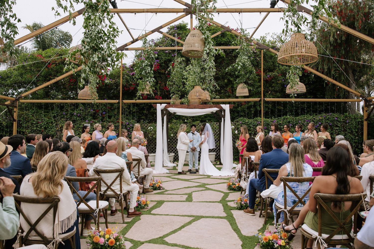 Outdoor wedding ceremony under bamboo structure with guests seated on wooden chairs.