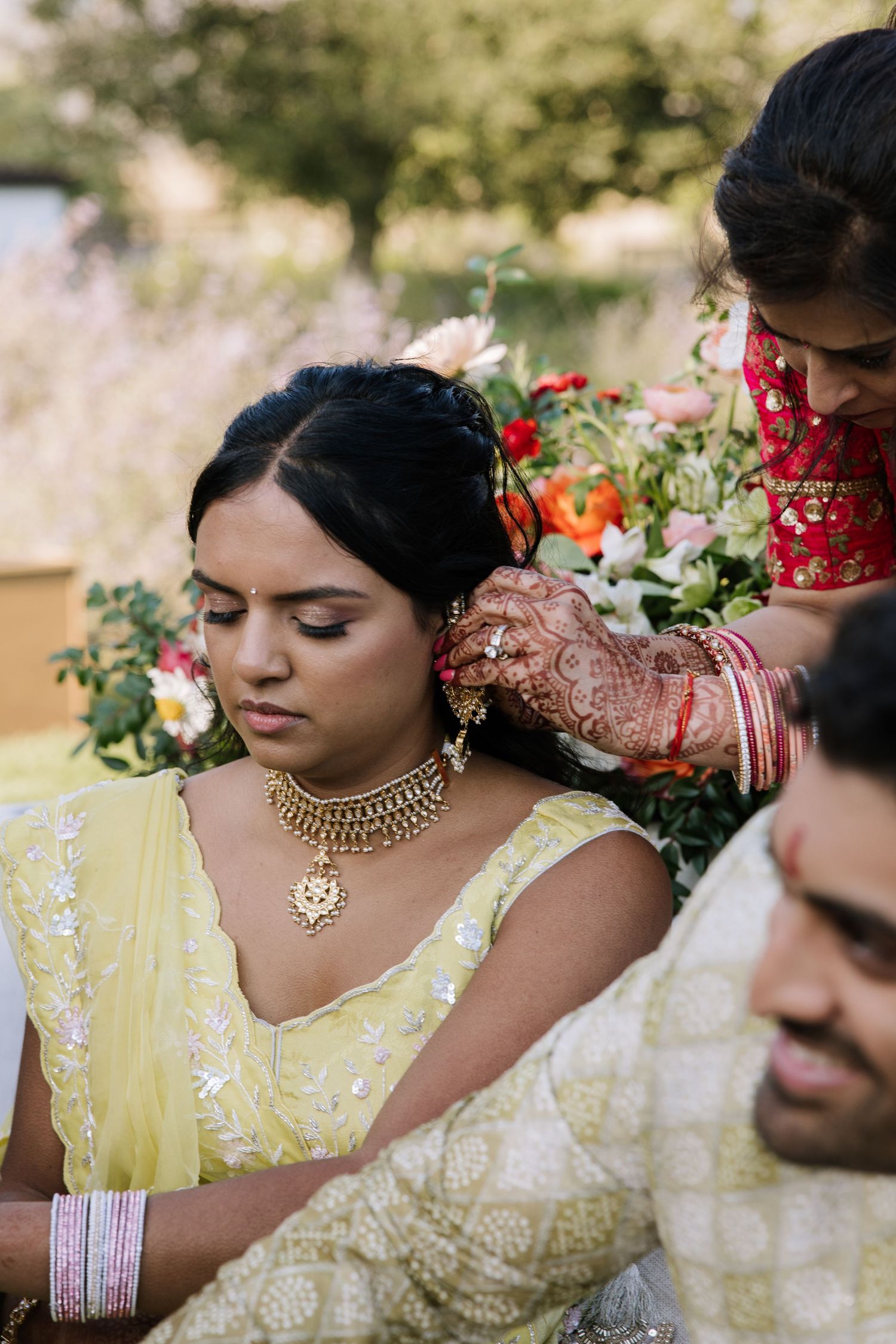 Indian bride in yellow lehenga with gold jewelry receives assistance during traditional outdoor wedding ceremony.
