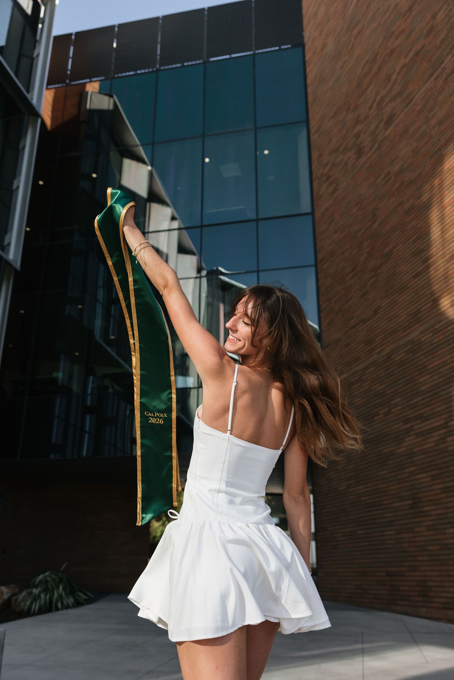 Young woman in white dress joyfully raising green graduation stole outside modern building.