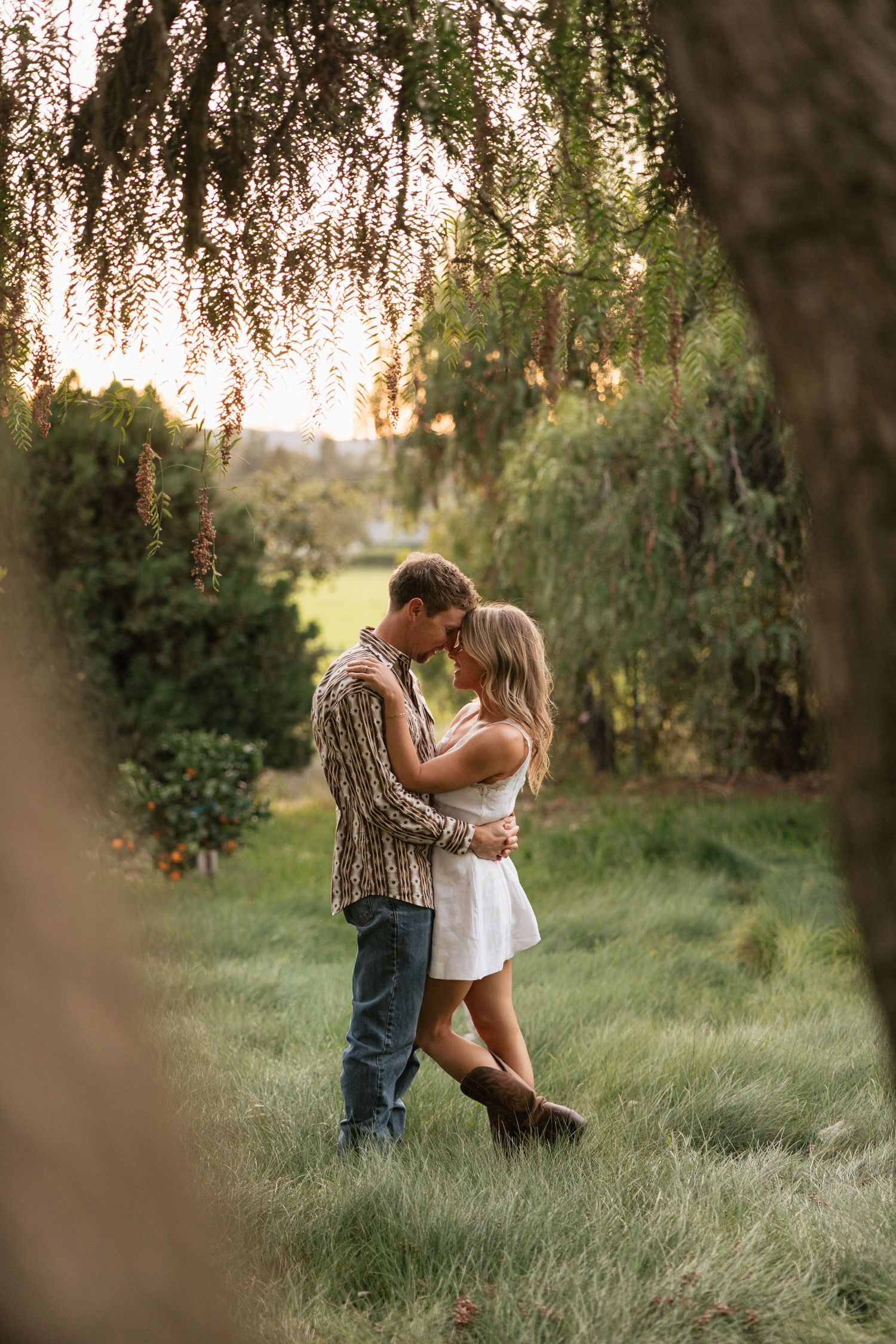 Romantic couple embracing under weeping willow branches in a lush green garden.