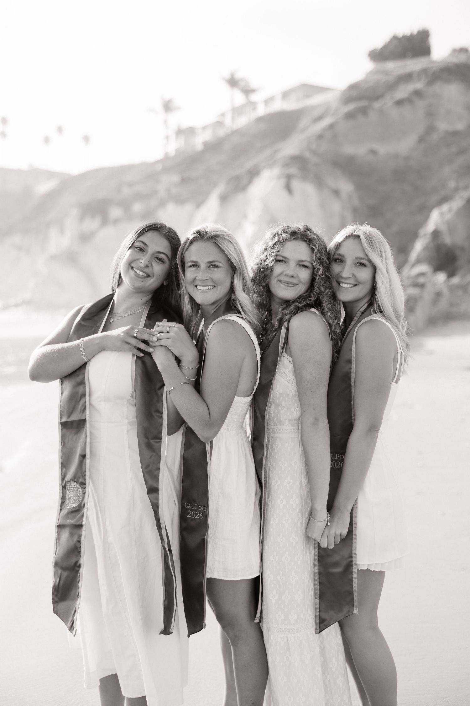 Four smiling female graduates in white dresses and graduation stoles pose on beach in black and white.