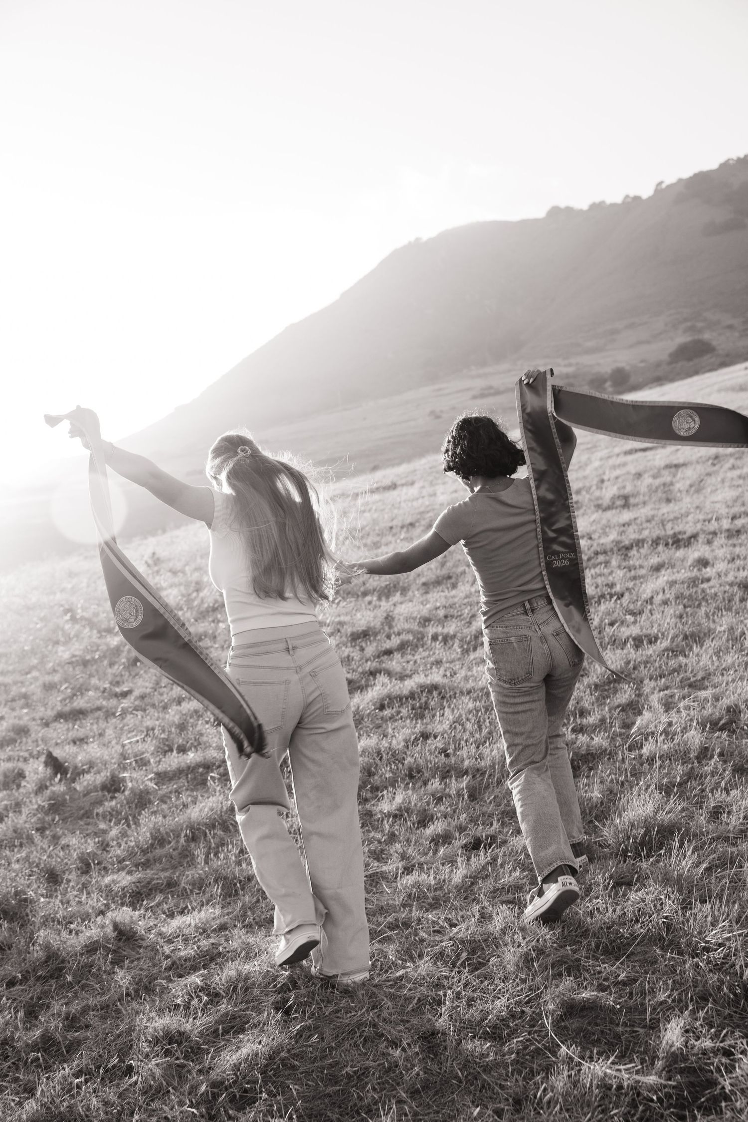 Black and white image of two women raising Cal Poly graduation sashes while walking through a sunlit field.