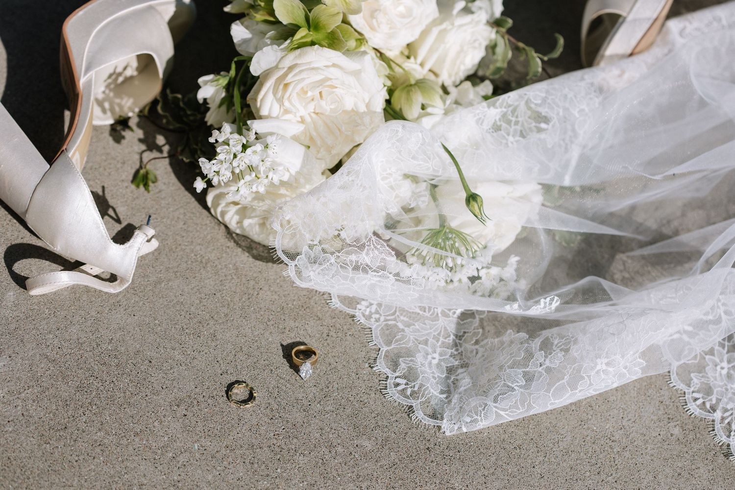 Wedding flat lay with white bouquet, lace veil, heels, and gold rings on gray surface.